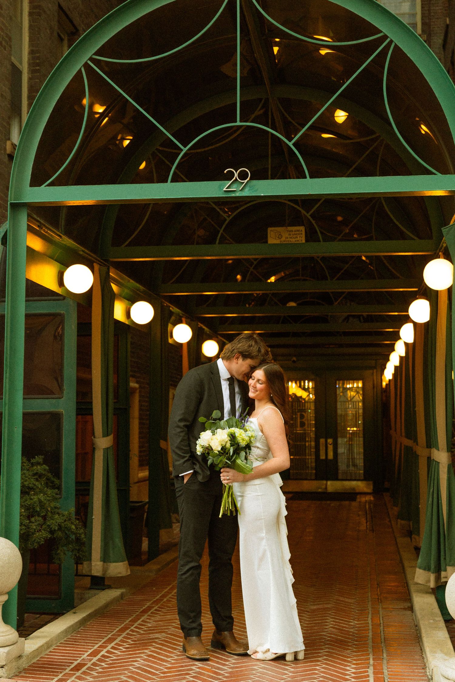 A couple stands in an elegant green archway with number 29, illuminated by a row of glowing pendant lights.