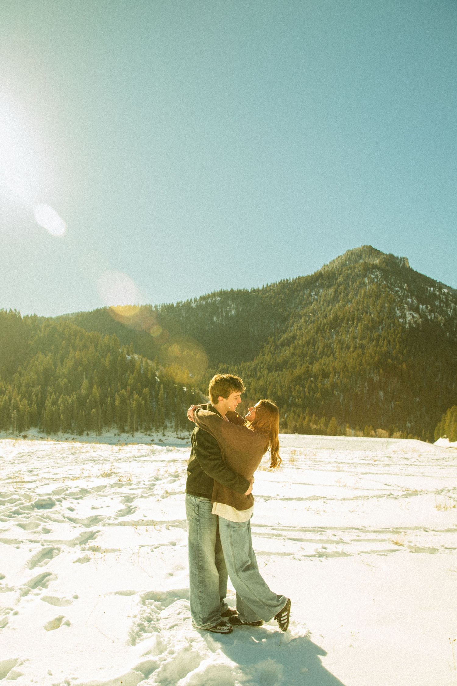 A couple embraces in the snow with sunlit mountain peaks in the background on a bright winter day.
