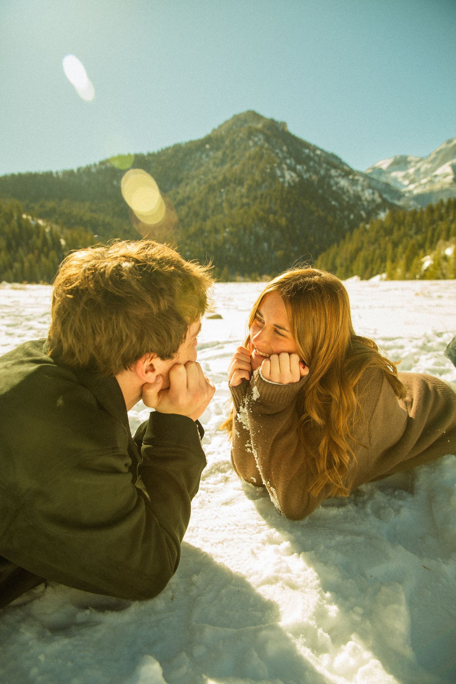 Two people playfully lying in snow with mountains visible in background during golden hour.