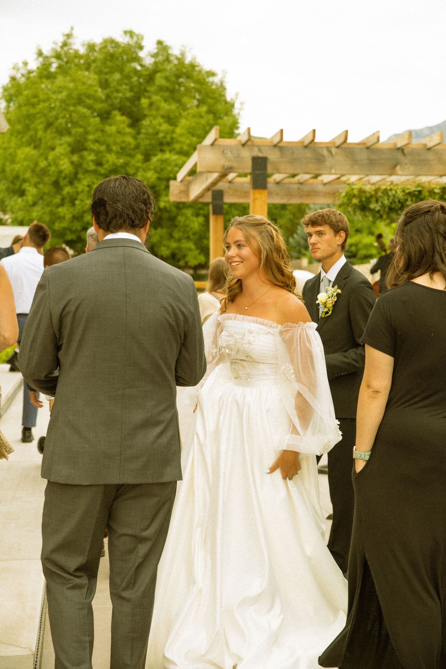 Wedding guests in formal attire gathered outdoors under a wooden pergola during a celebration.