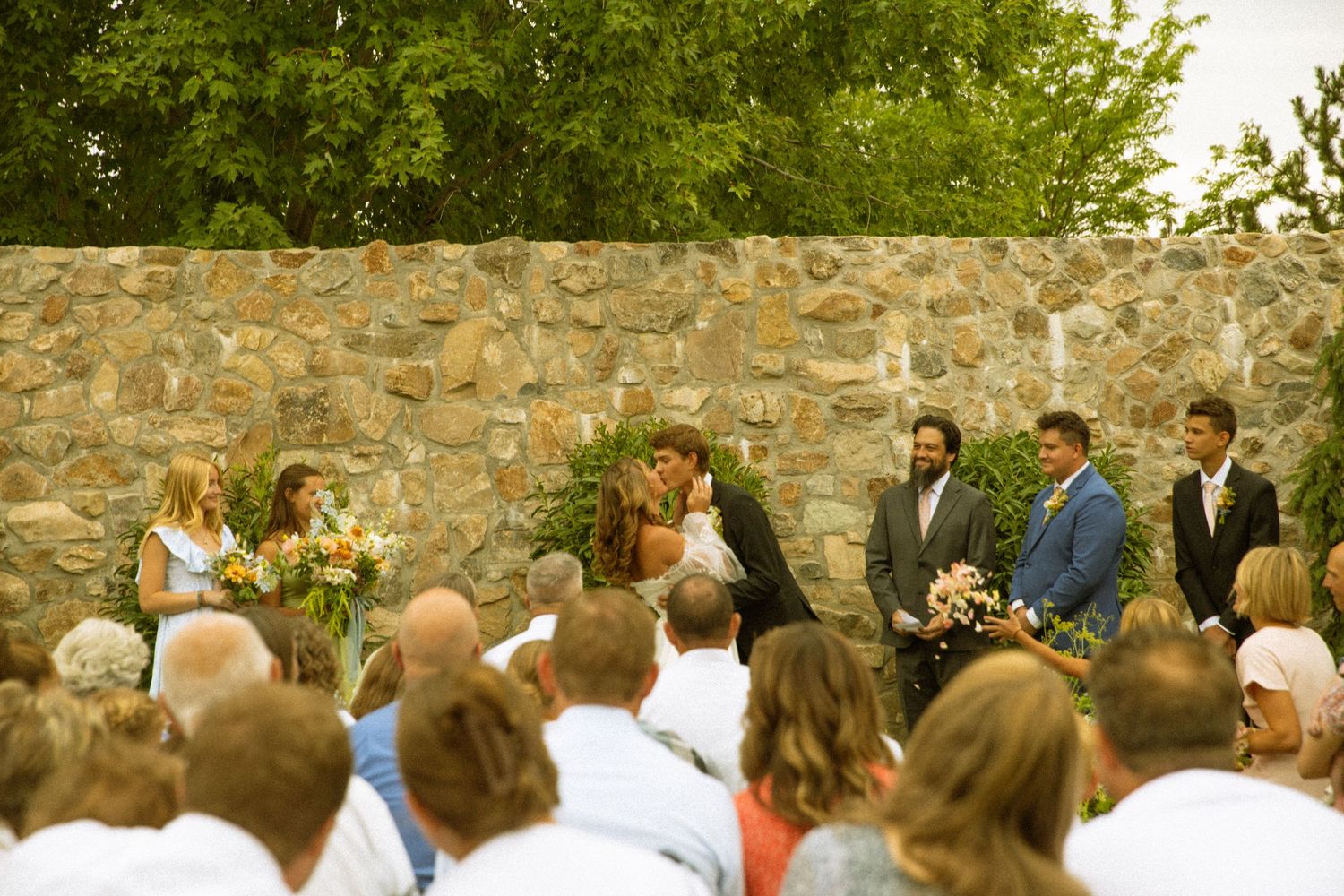 Wedding ceremony attendees gathered outdoors by stone wall with flowering garden.