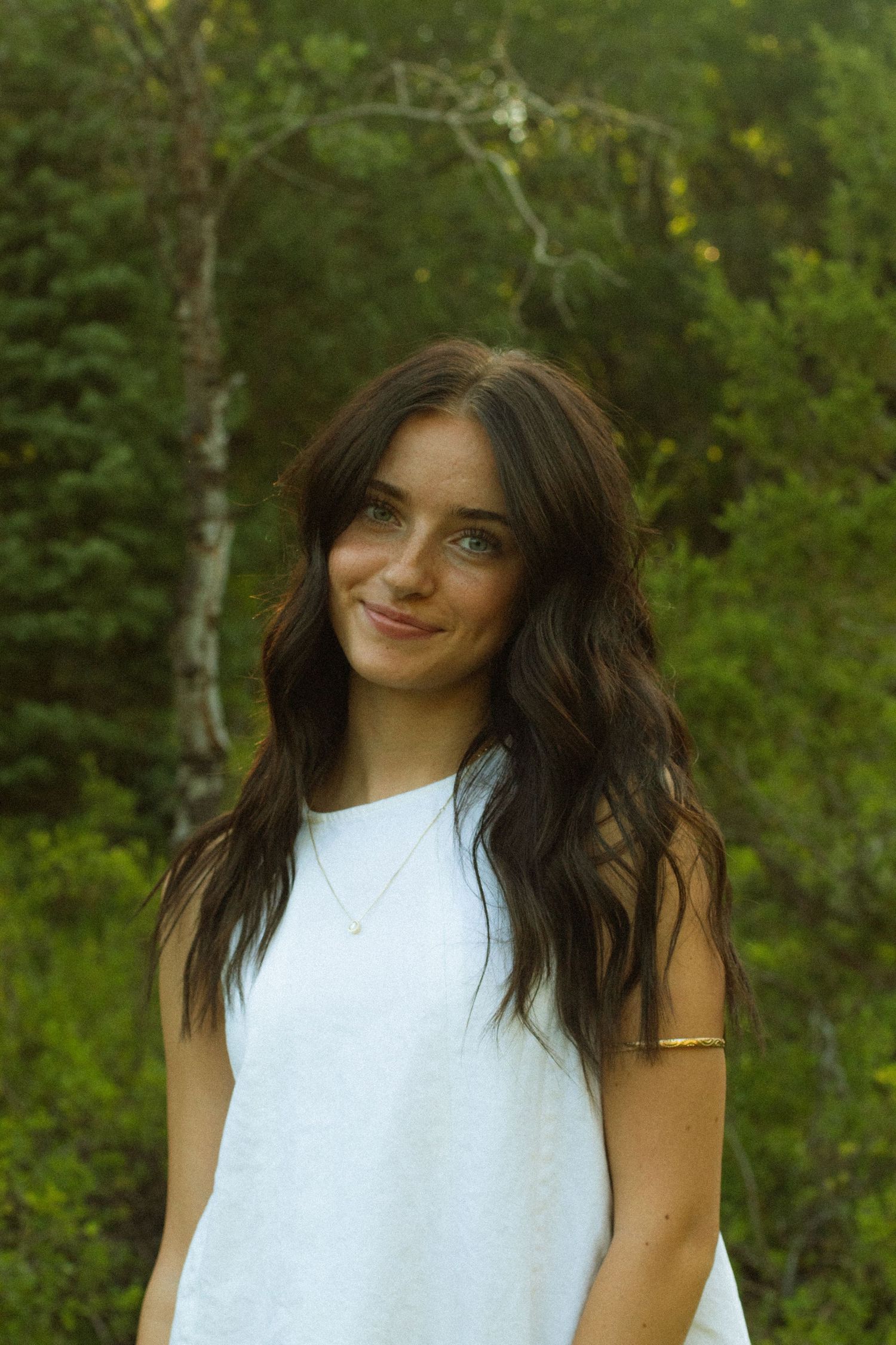 A person in a white sleeveless top poses playfully against a lush green forest background.