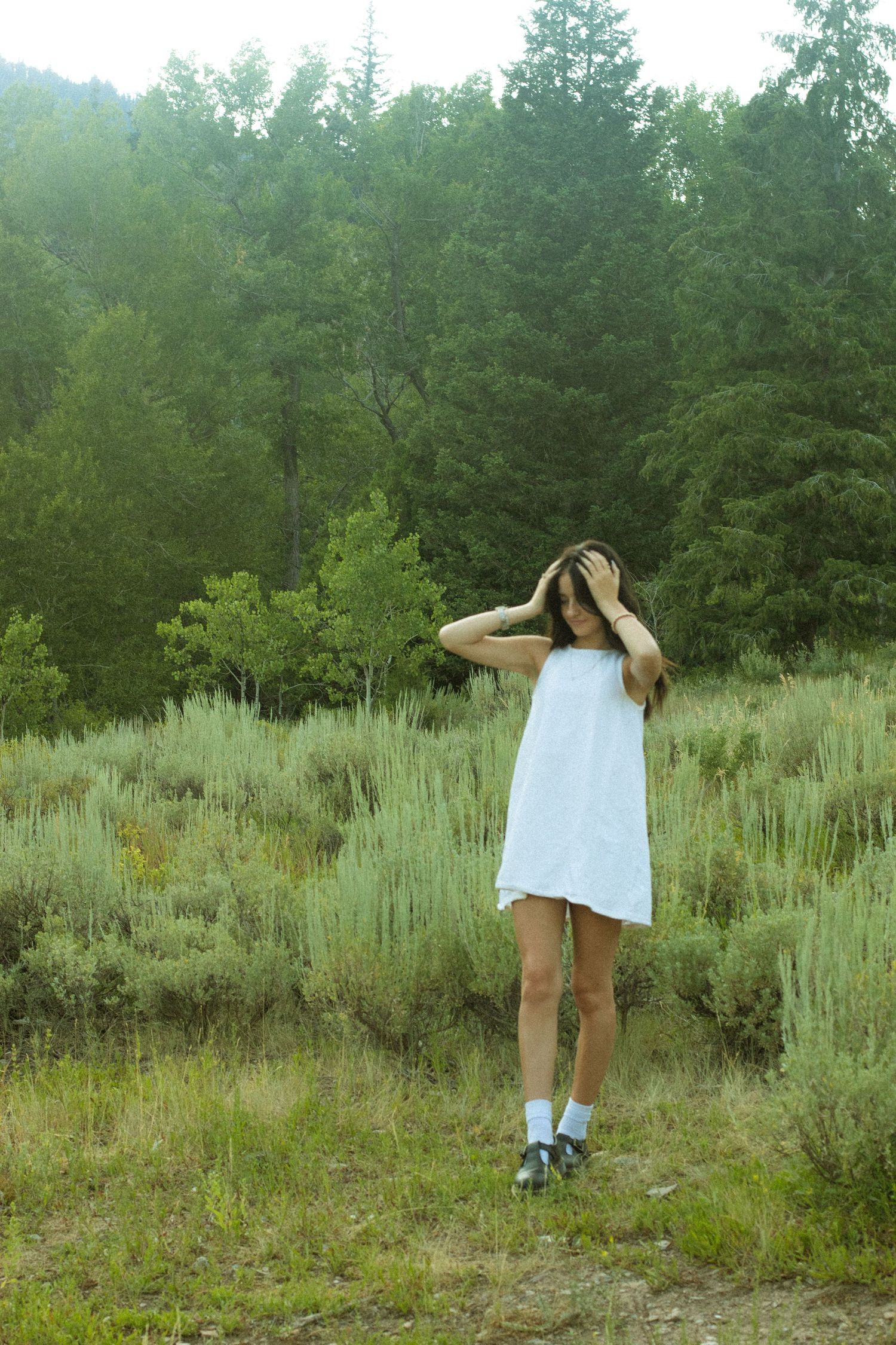 A person in a white dress stands among sagebrush and evergreen trees in a misty natural landscape.