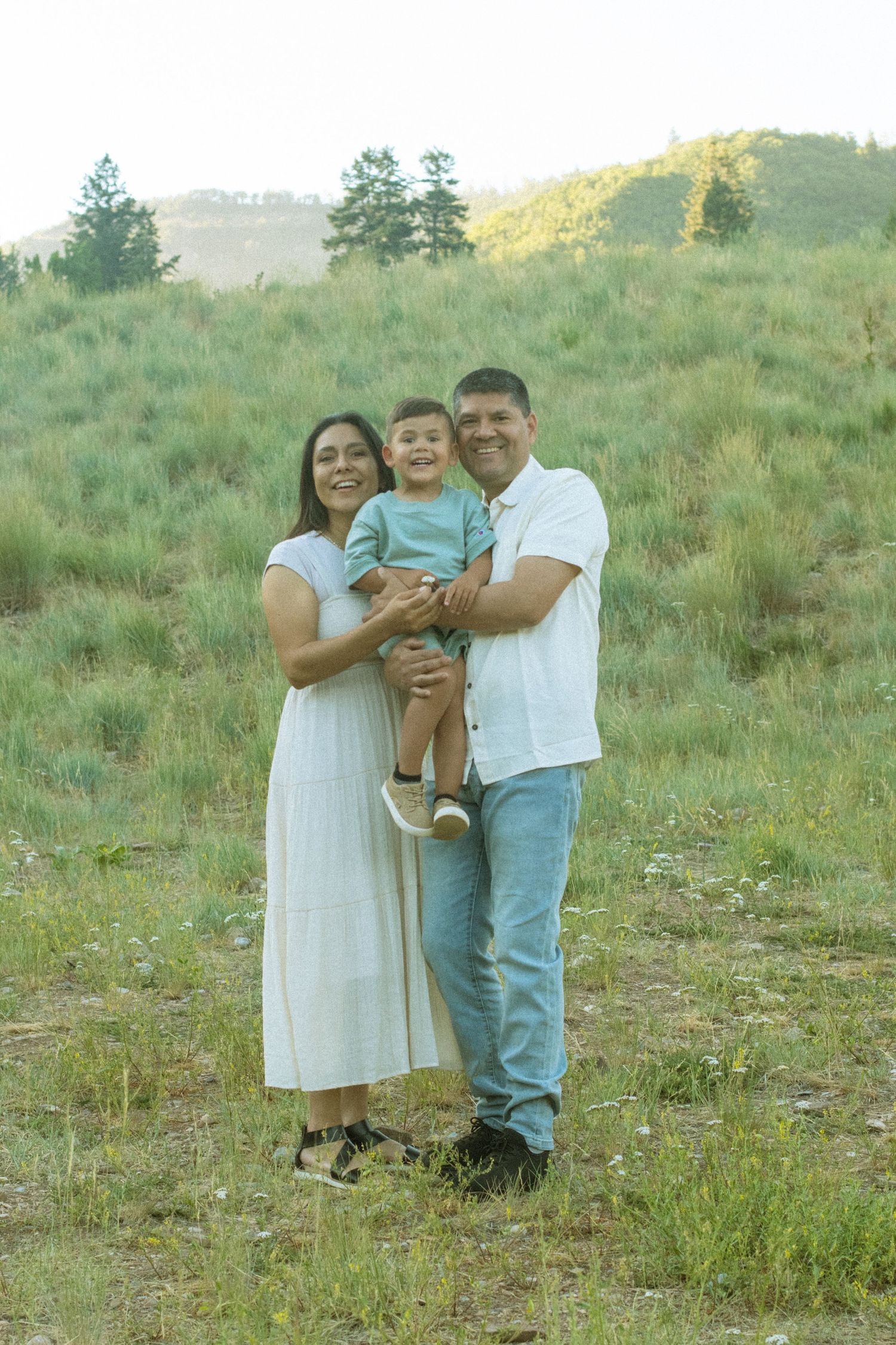 A family of three poses together on a sunny hillside with mountains visible in the distance.