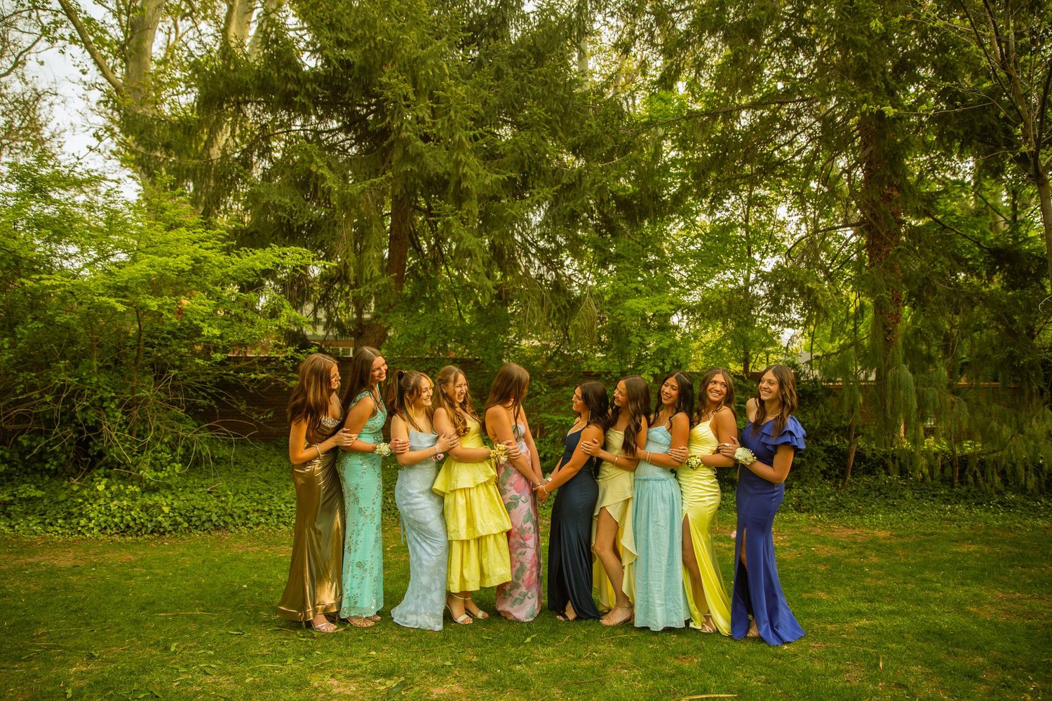 A group of bridesmaids in colorful mismatched gowns laughing together on a lush green lawn surrounded by tall trees.