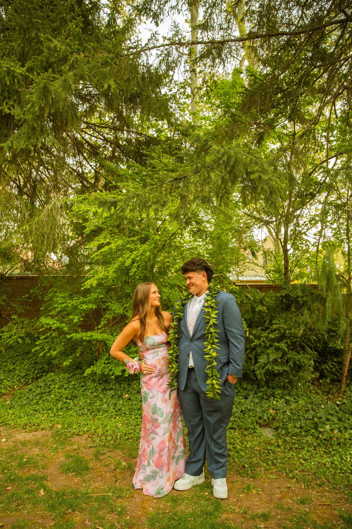 A young couple in formal prom attire poses outdoors among lush green trees, wearing floral lei garlands.