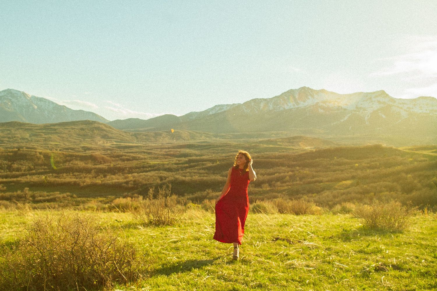 Woman in red dress standing in a sunlit mountain valley landscape at golden hour.
