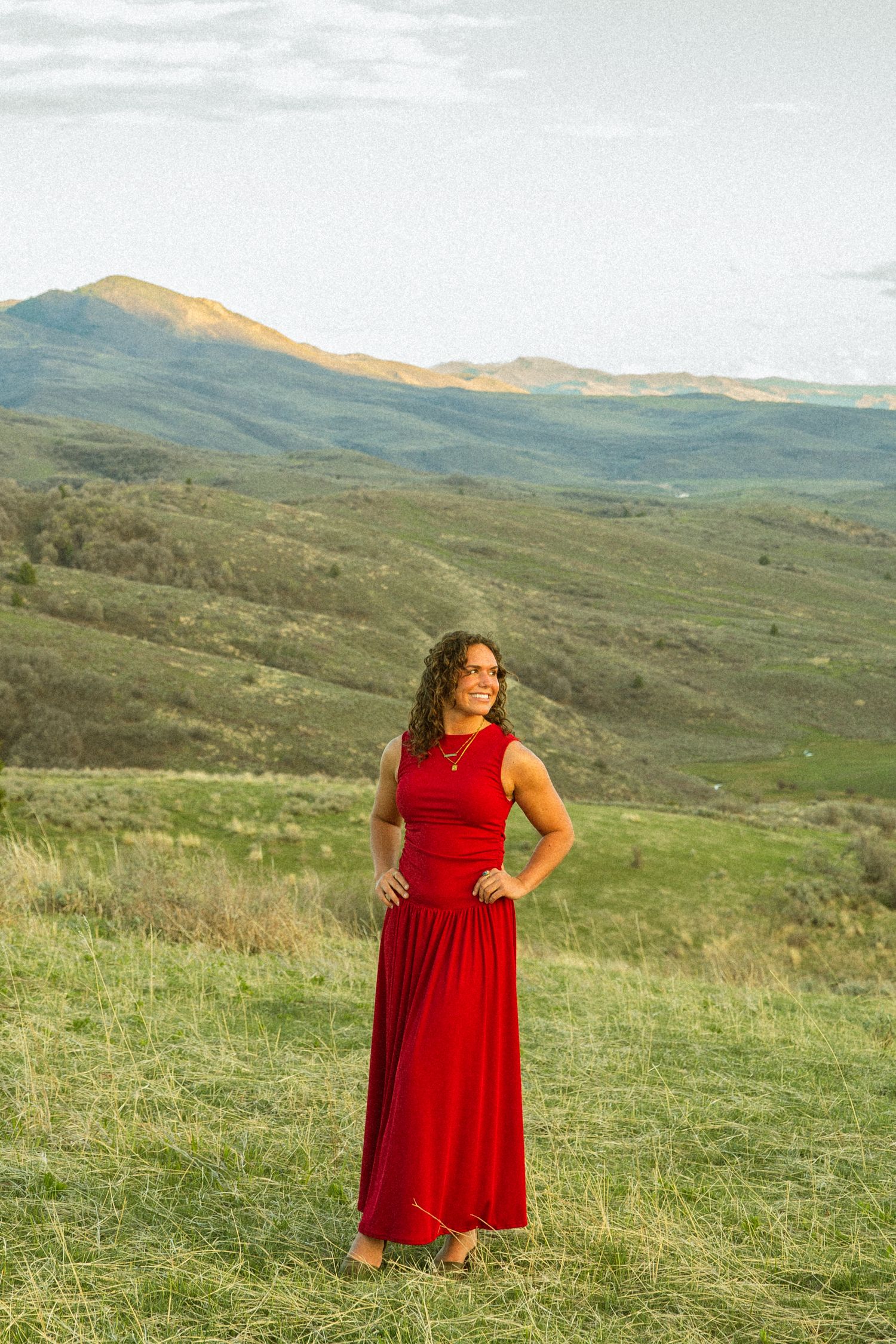 Woman in red maxi dress standing on green hillside with mountain landscape backdrop in golden hour light.