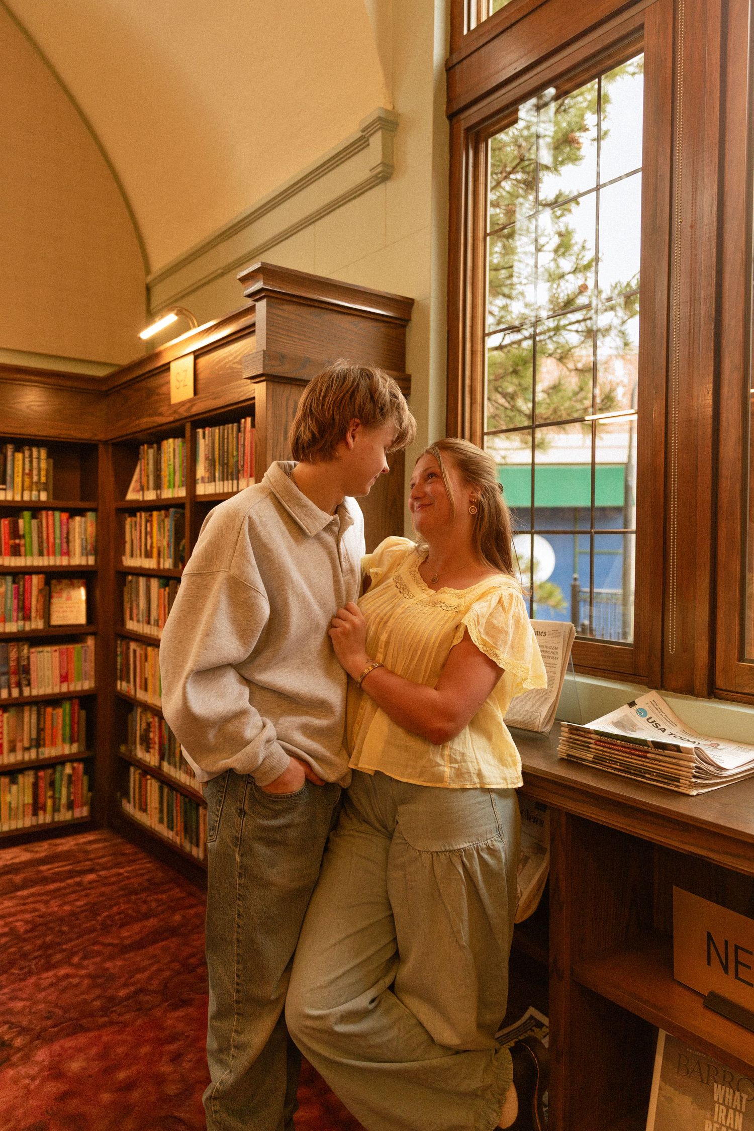 A couple embraces while browsing books in a cozy library with wooden shelves and red carpeting.