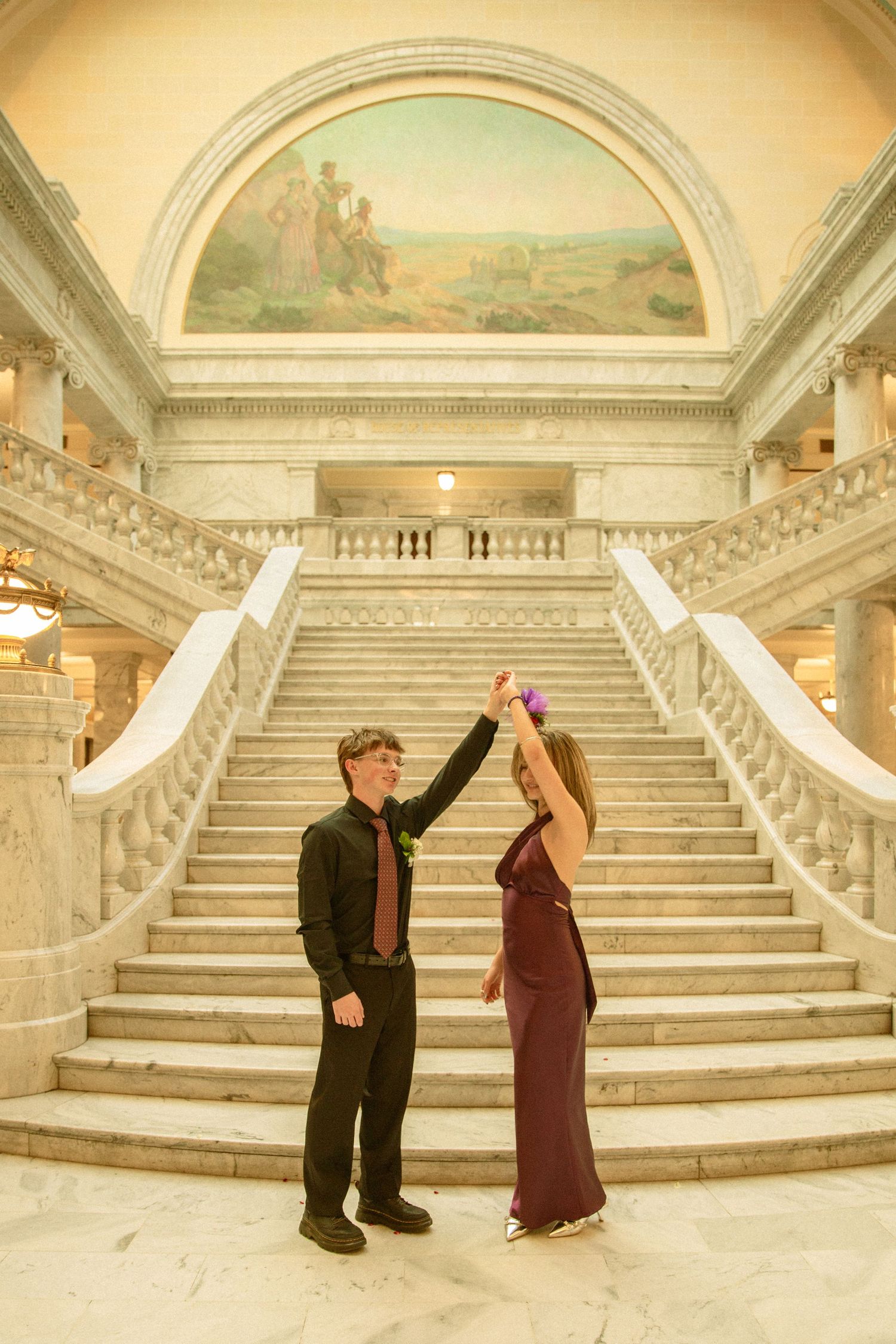 Elegant couple dancing on marble staircase in grand historic building with arched ceiling and mural above.