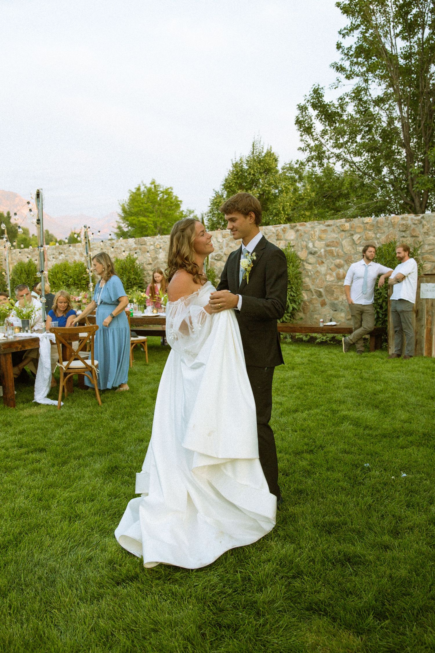 Wedding guests dancing on a grassy lawn near a stone wall during an outdoor celebration.