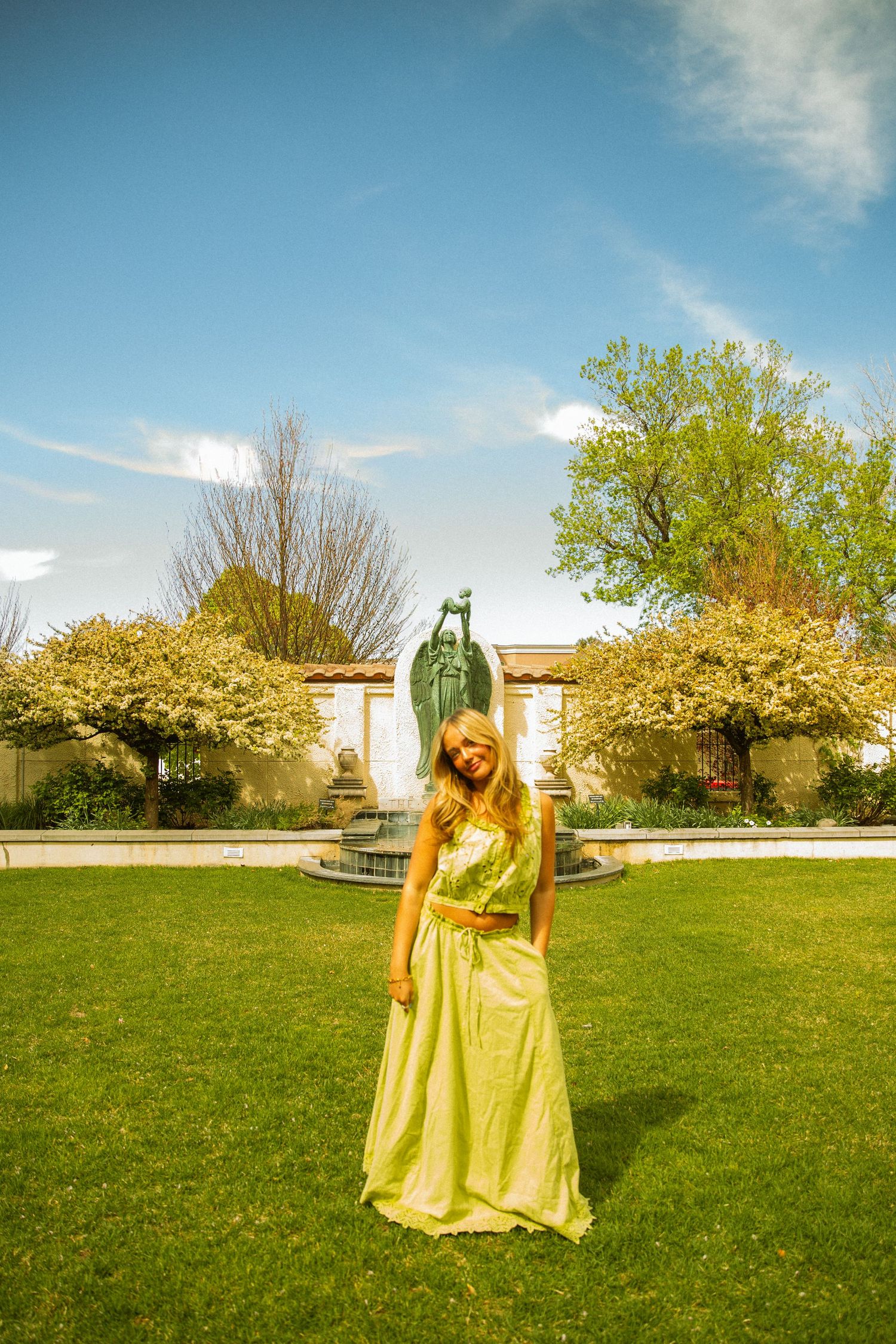 Woman in yellow gown poses on green lawn near bronze fountain sculpture with flowering trees and blue sky.