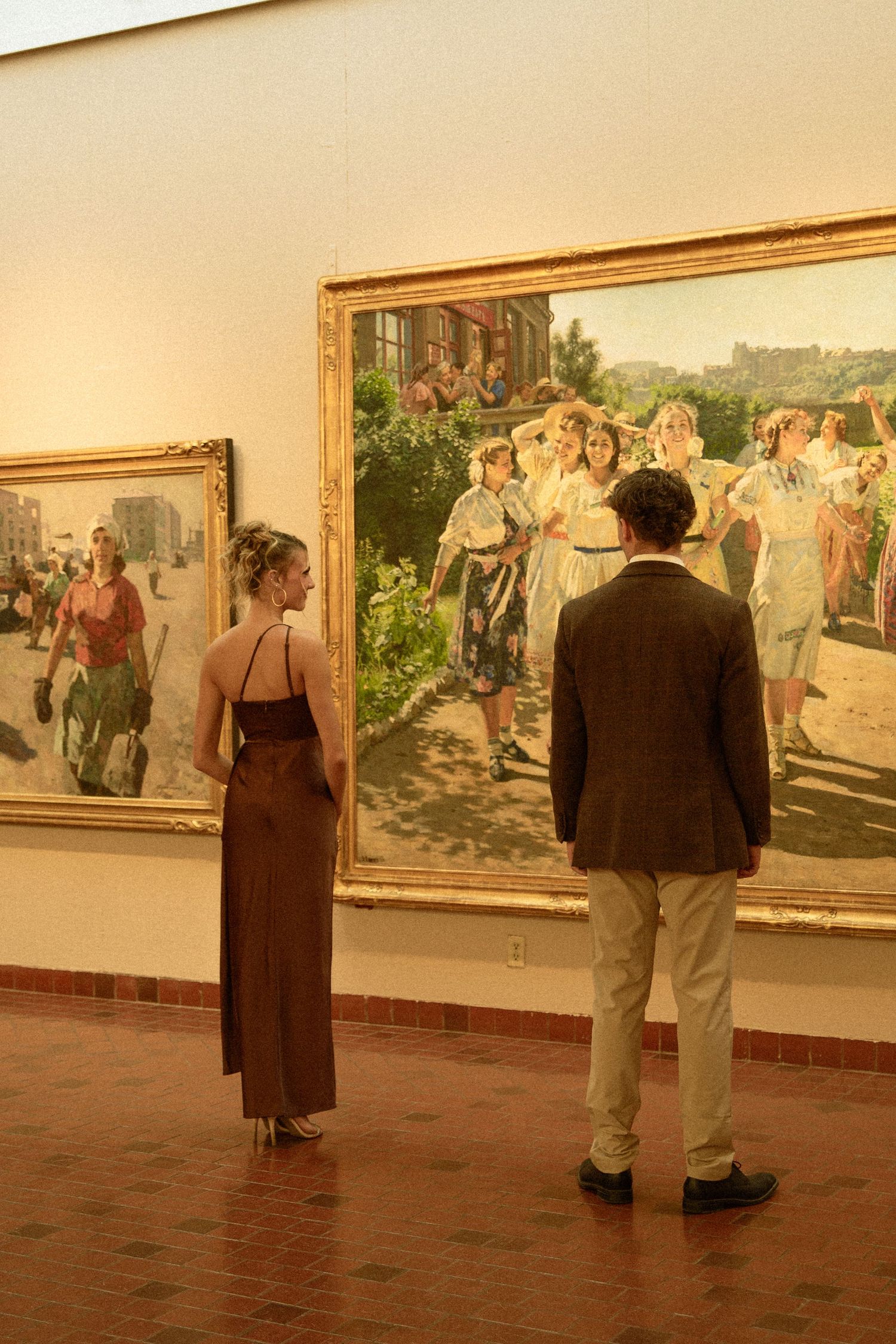 Museum visitors view large paintings displayed on gallery walls with terracotta flooring in warm lighting.