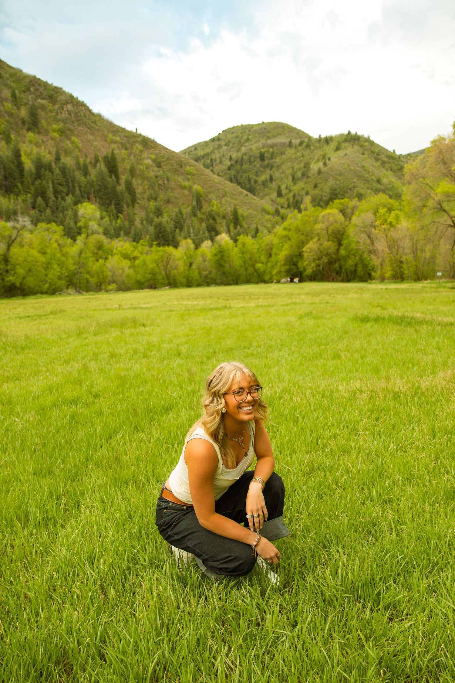 Smiling blonde woman with glasses crouching in a lush green meadow with forested mountains in the background.