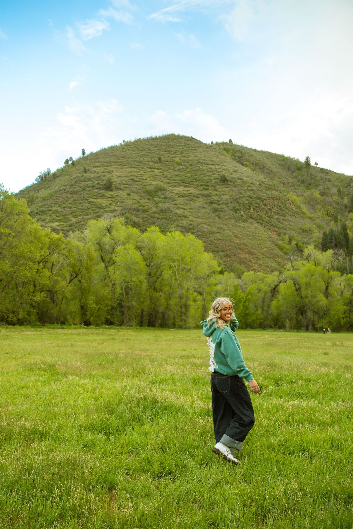 A blonde woman in a green hoodie walks through a lush meadow with a tree-covered hill and blue sky behind her.