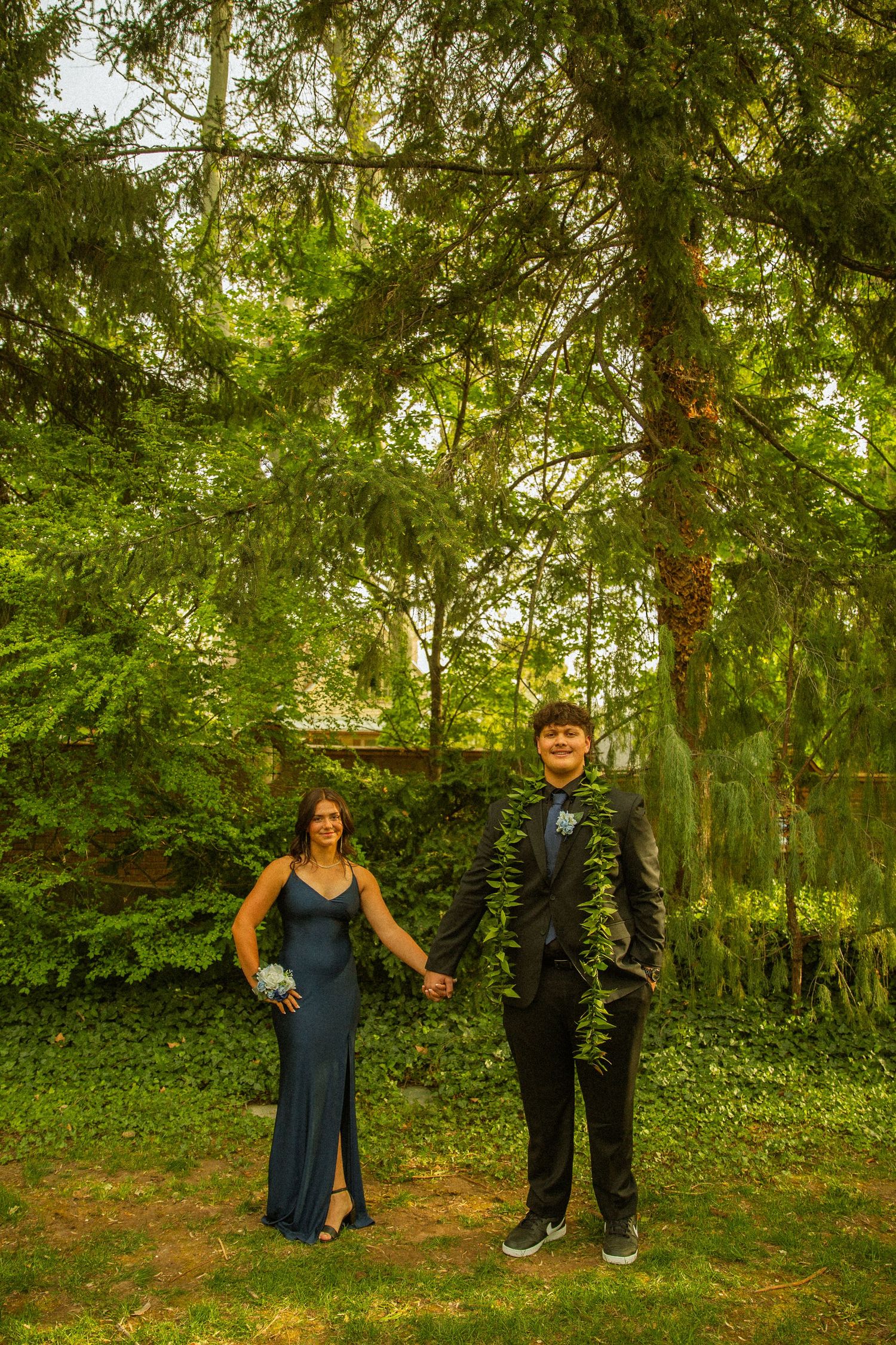 A couple in prom attire, woman in blue gown and man in black suit with lei, pose holding hands outdoors.