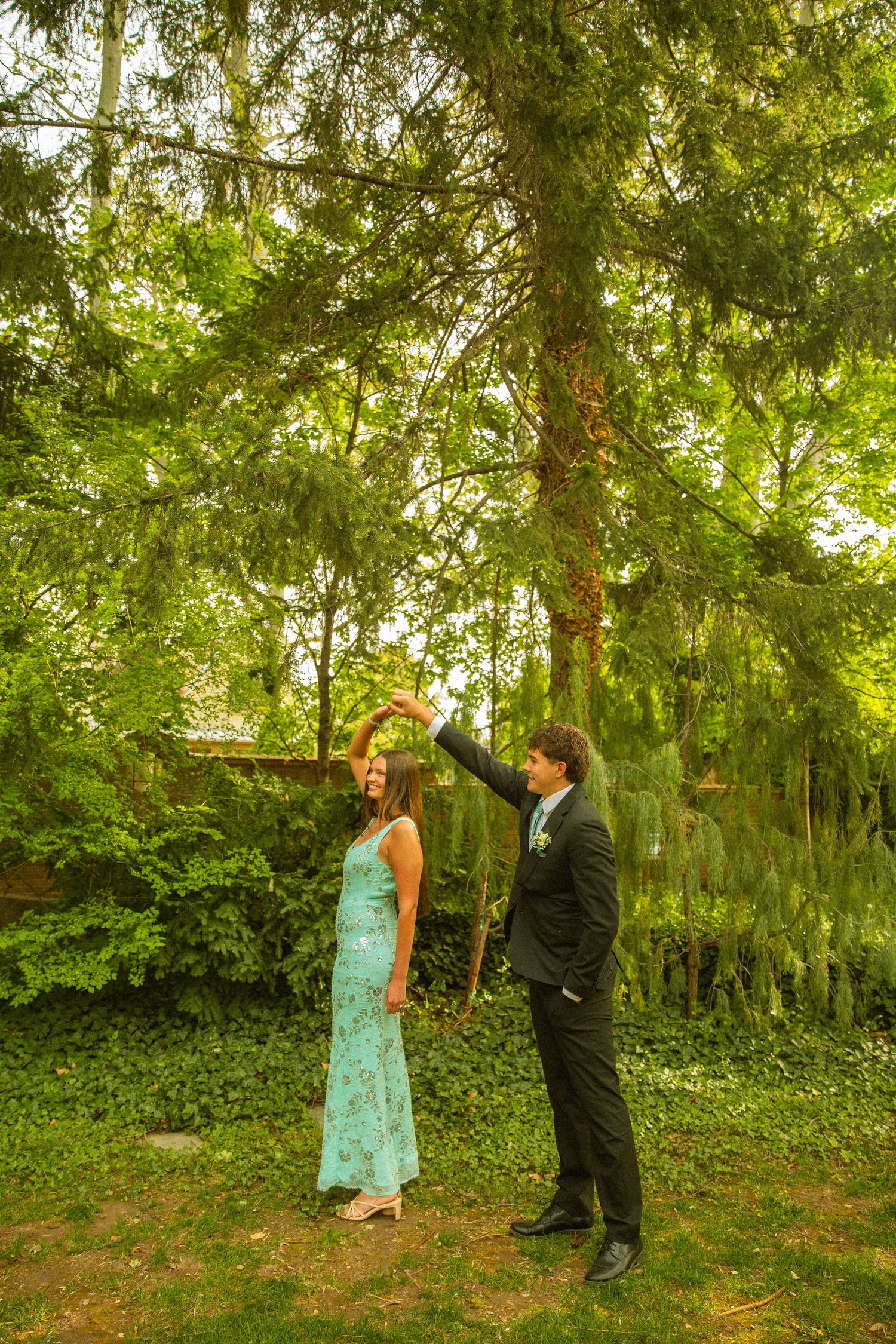 Couple dancing outdoors in lush green garden, woman in teal gown, man in black suit spinning her under tall trees.
