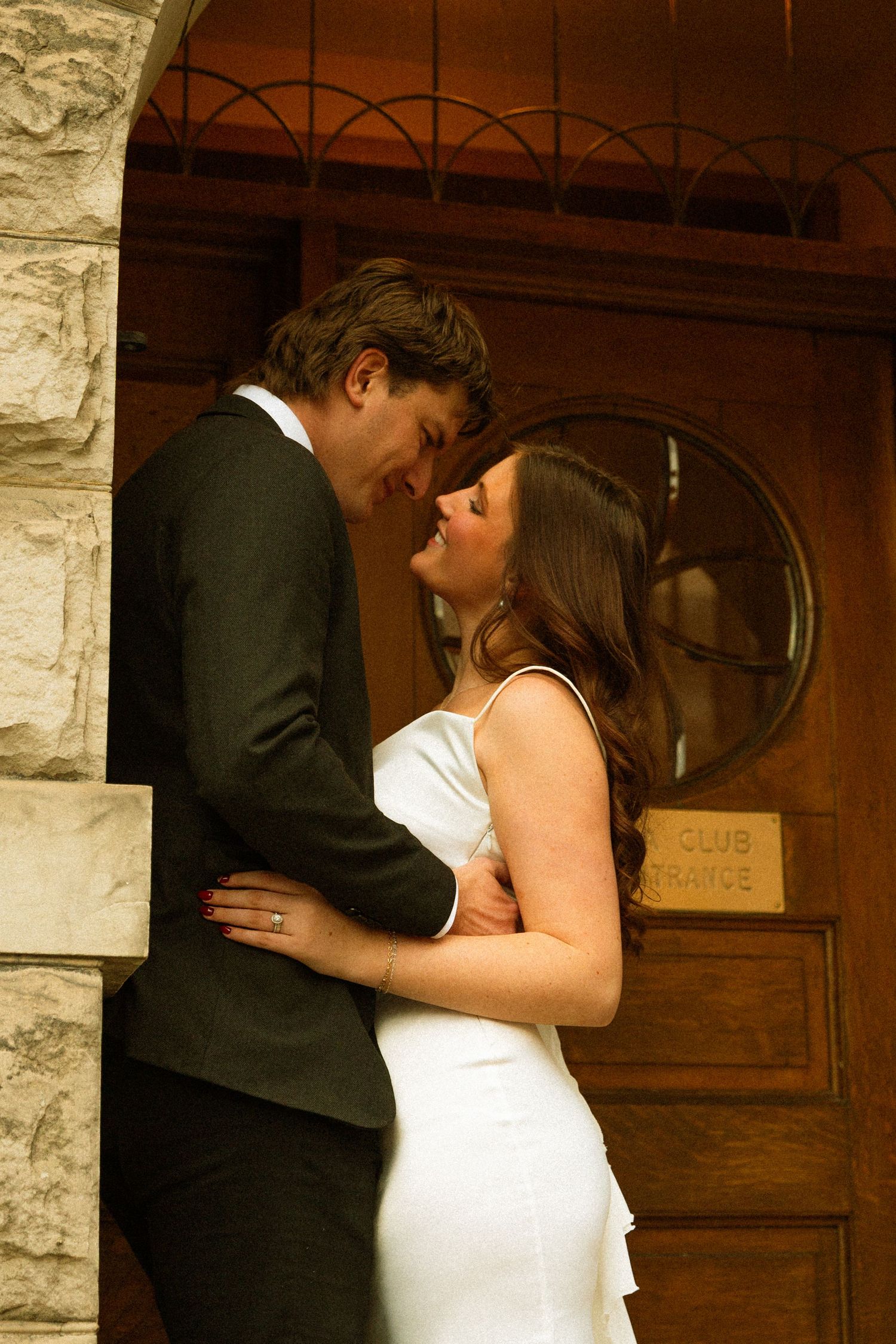 A romantic couple shares an intimate moment in front of a rustic wooden door with stone walls.