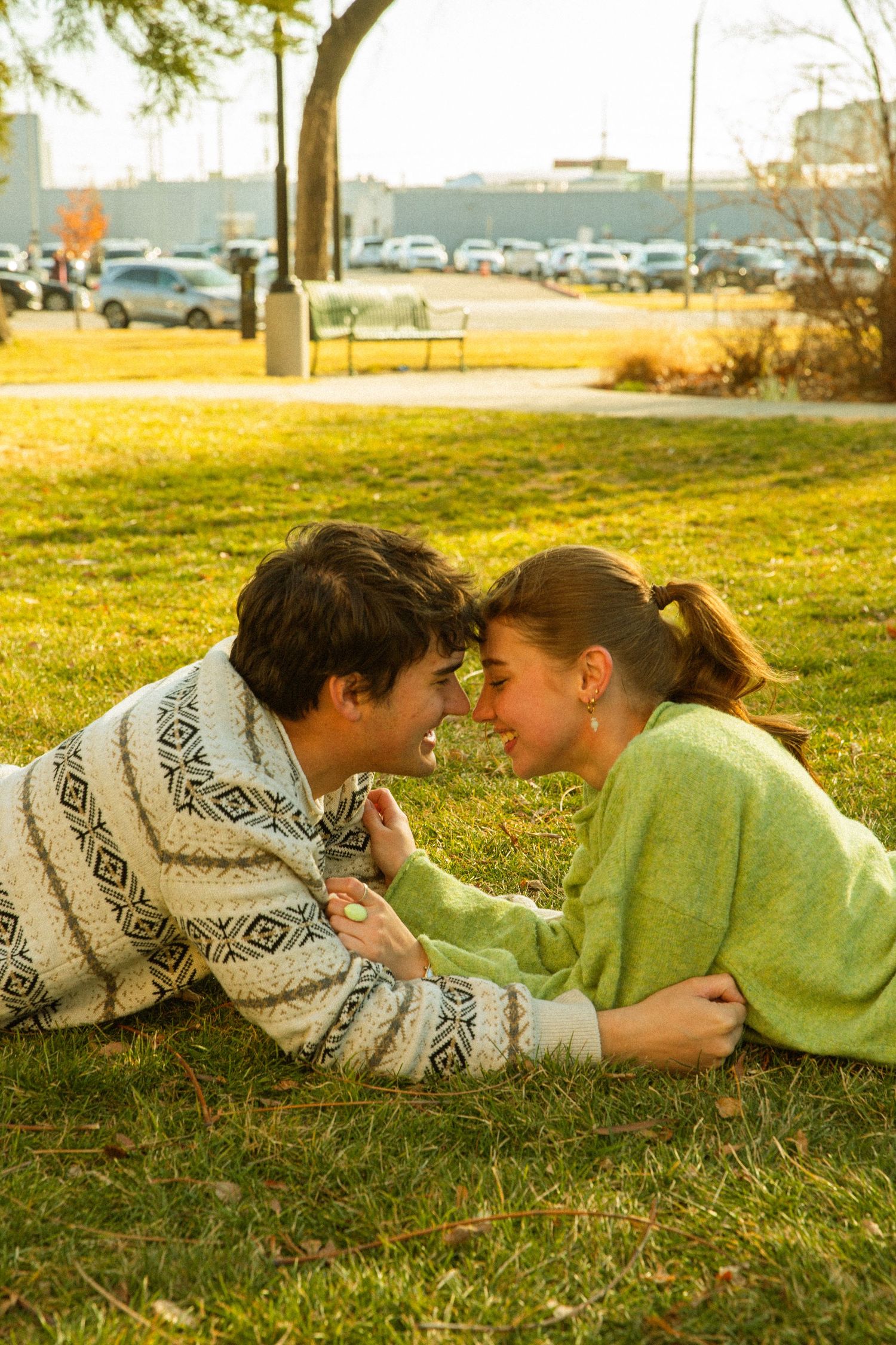A couple in cozy sweaters lying on grass in a park during golden hour with cars and trees in background.