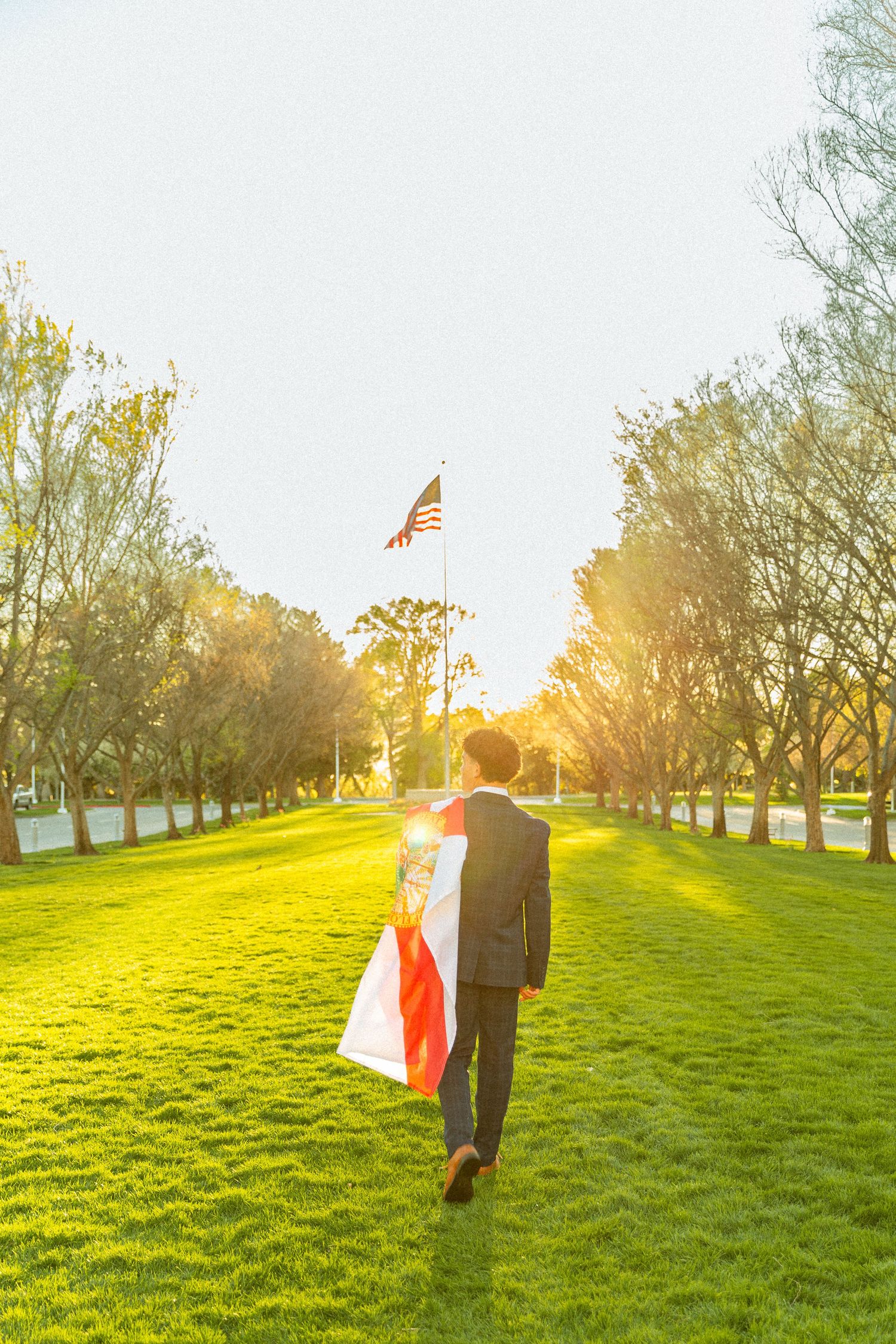 Man in suit walking toward American flag on sunny green lawn lined with trees.