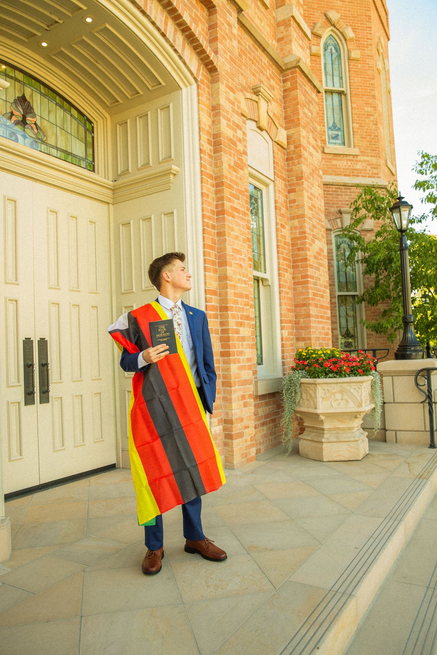 A graduate in colorful academic regalia stands outside a brick building with arched doorways and decorative windows.
