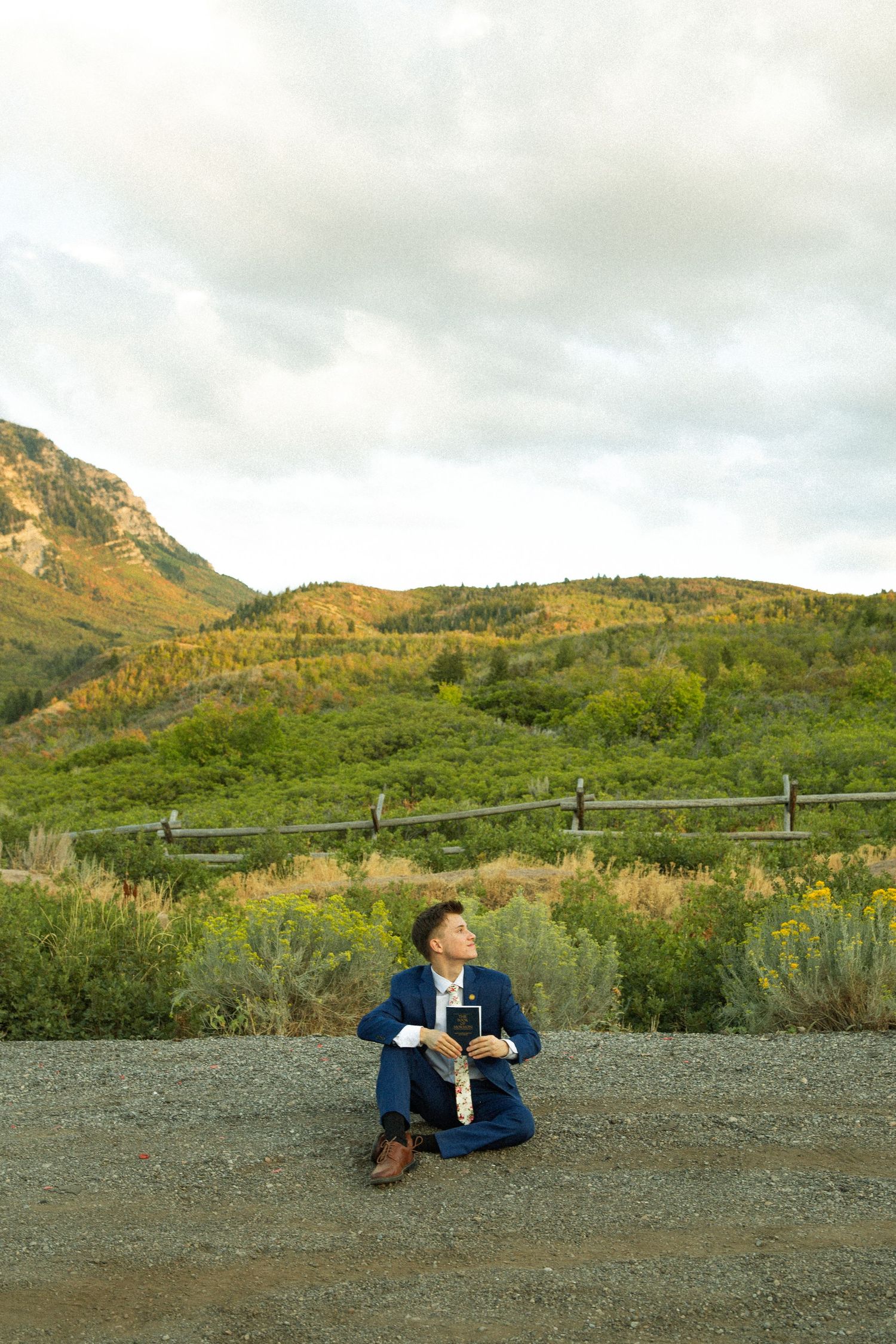 A person in a blue jacket sits on a gravel path against a backdrop of green mountains and cloudy skies.