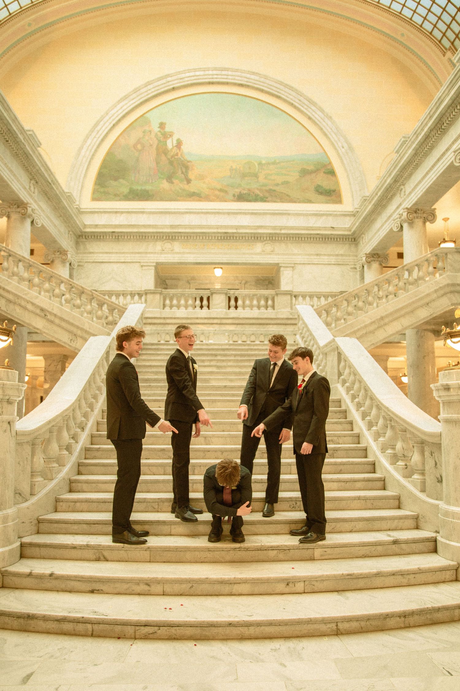 A group stands on an elegant marble staircase with ornate architecture and a mural in the background.