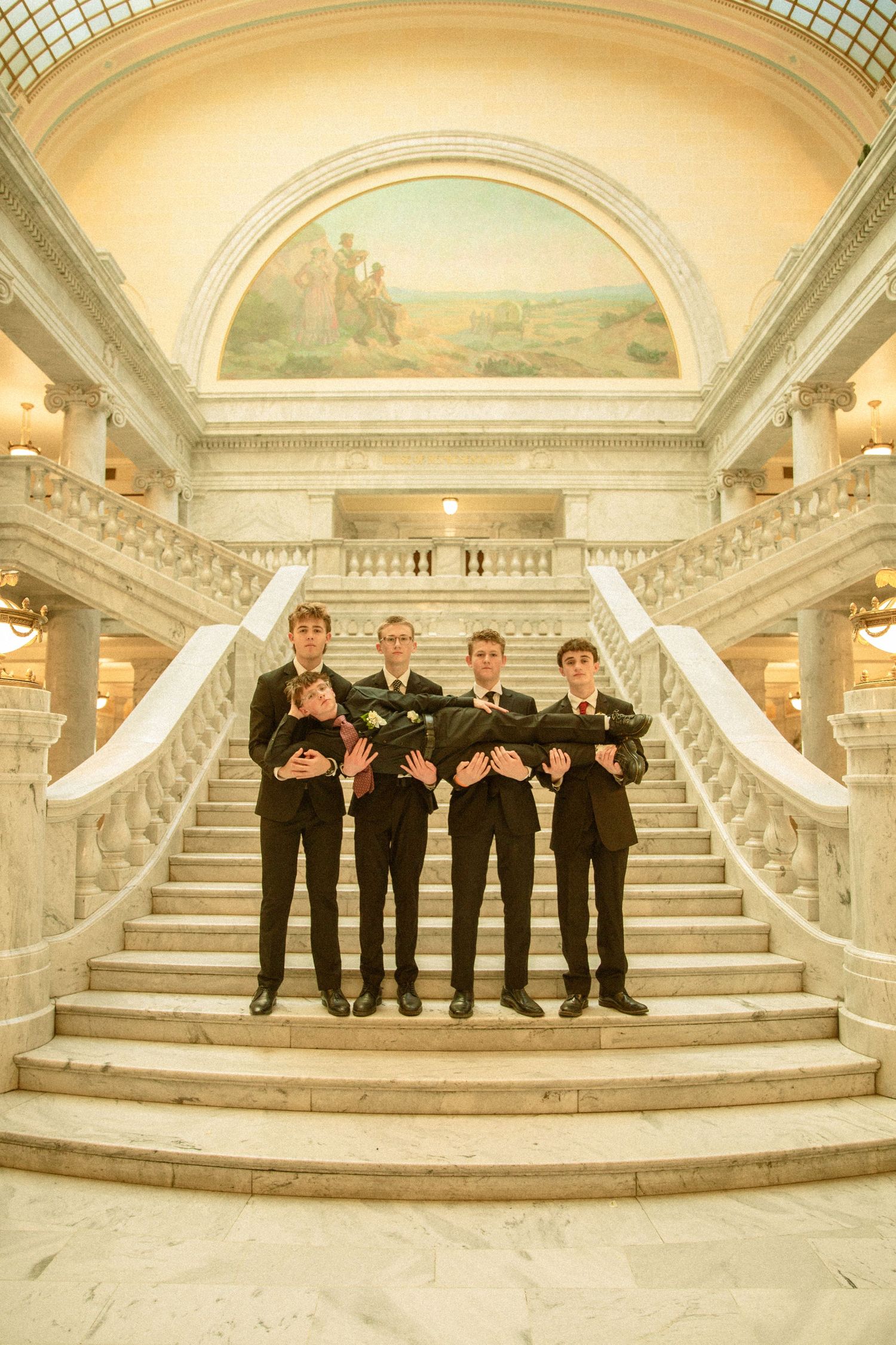 Four groomsmen in matching black suits stand on an elegant marble staircase in a grand historic building interior.