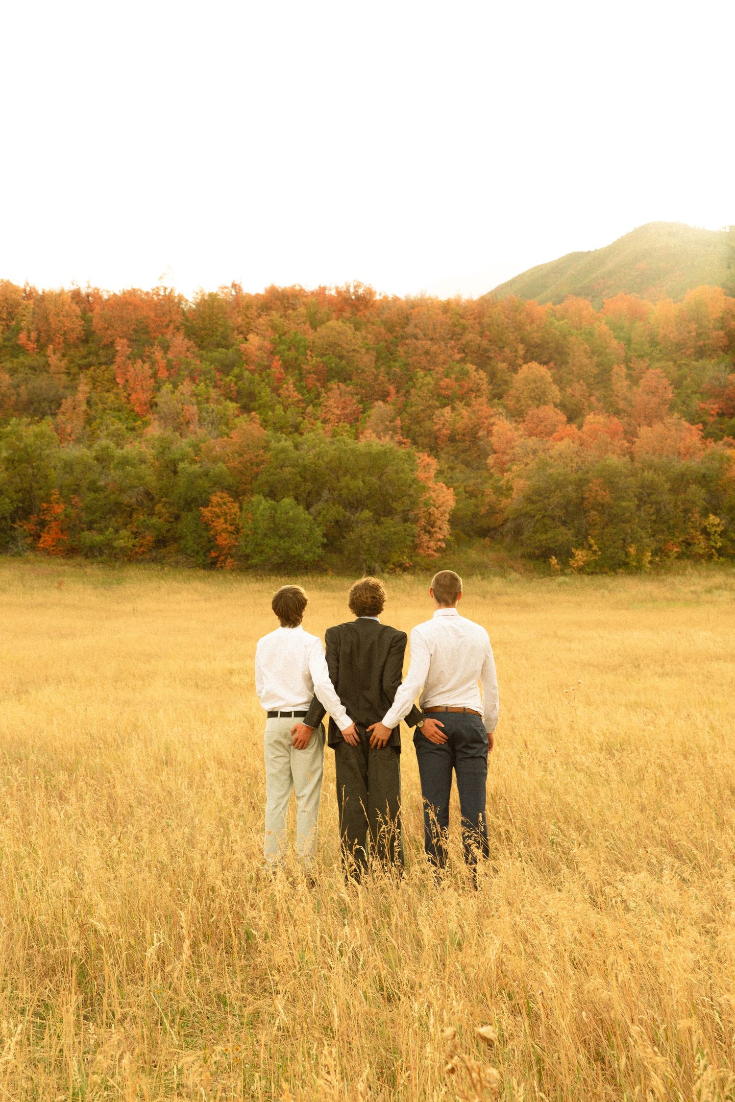 Three people stand together in a golden field with autumn trees on the hillside during sunset.