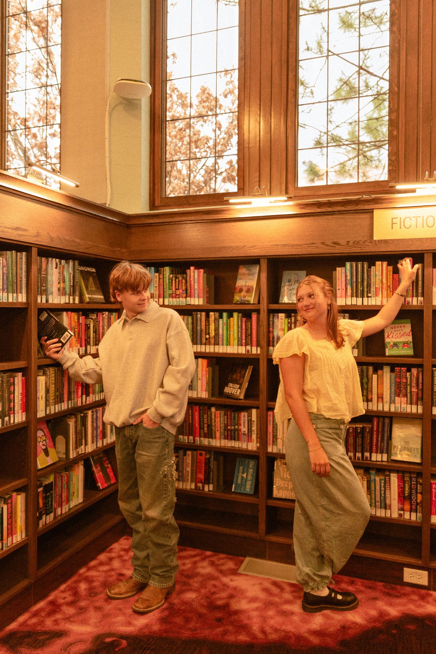 Two young people posing casually between bookshelves in a warm-lit library with wooden trim and tall windows.