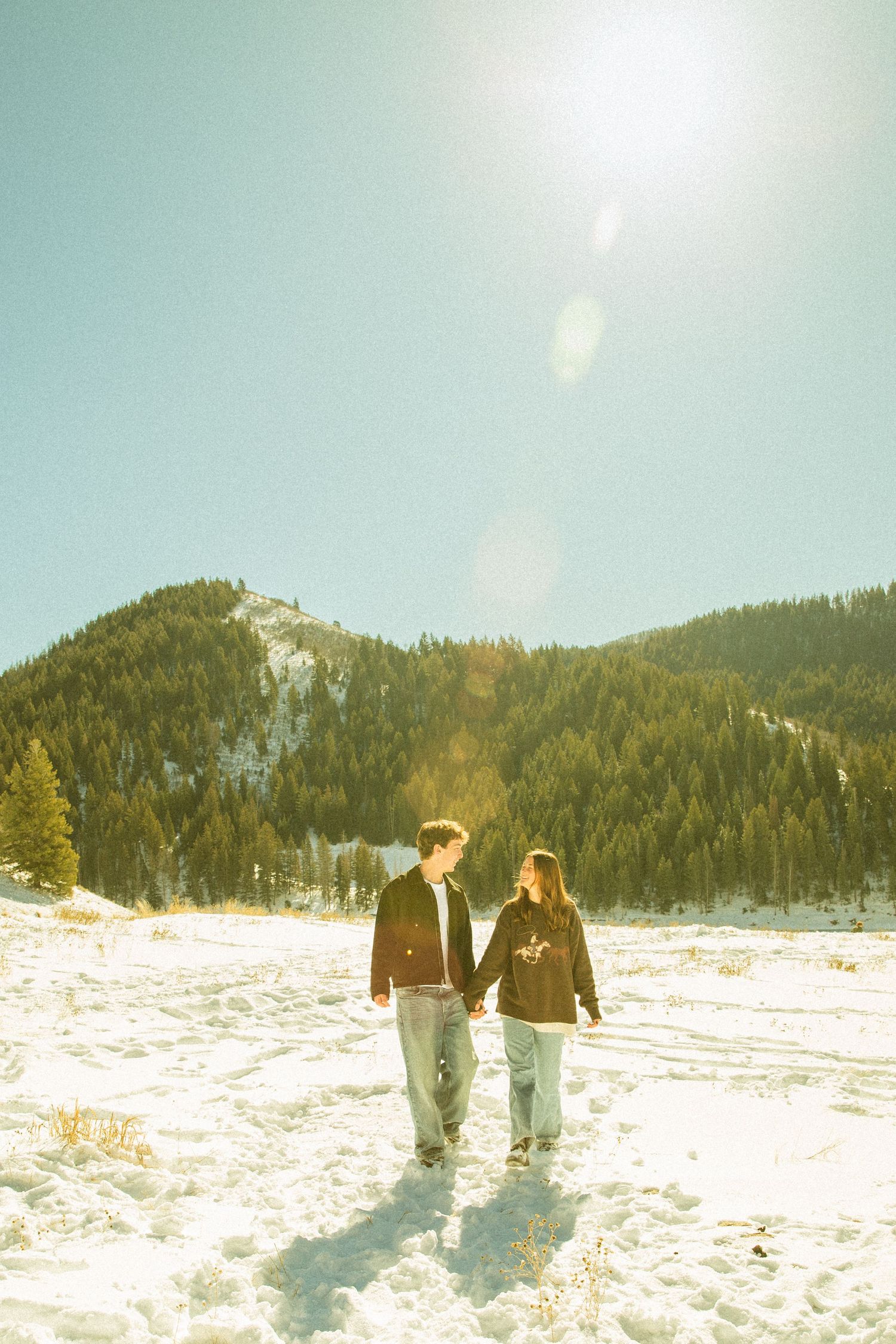 Two people walk together across a snowy mountain landscape on a bright winter day with evergreen trees in background.