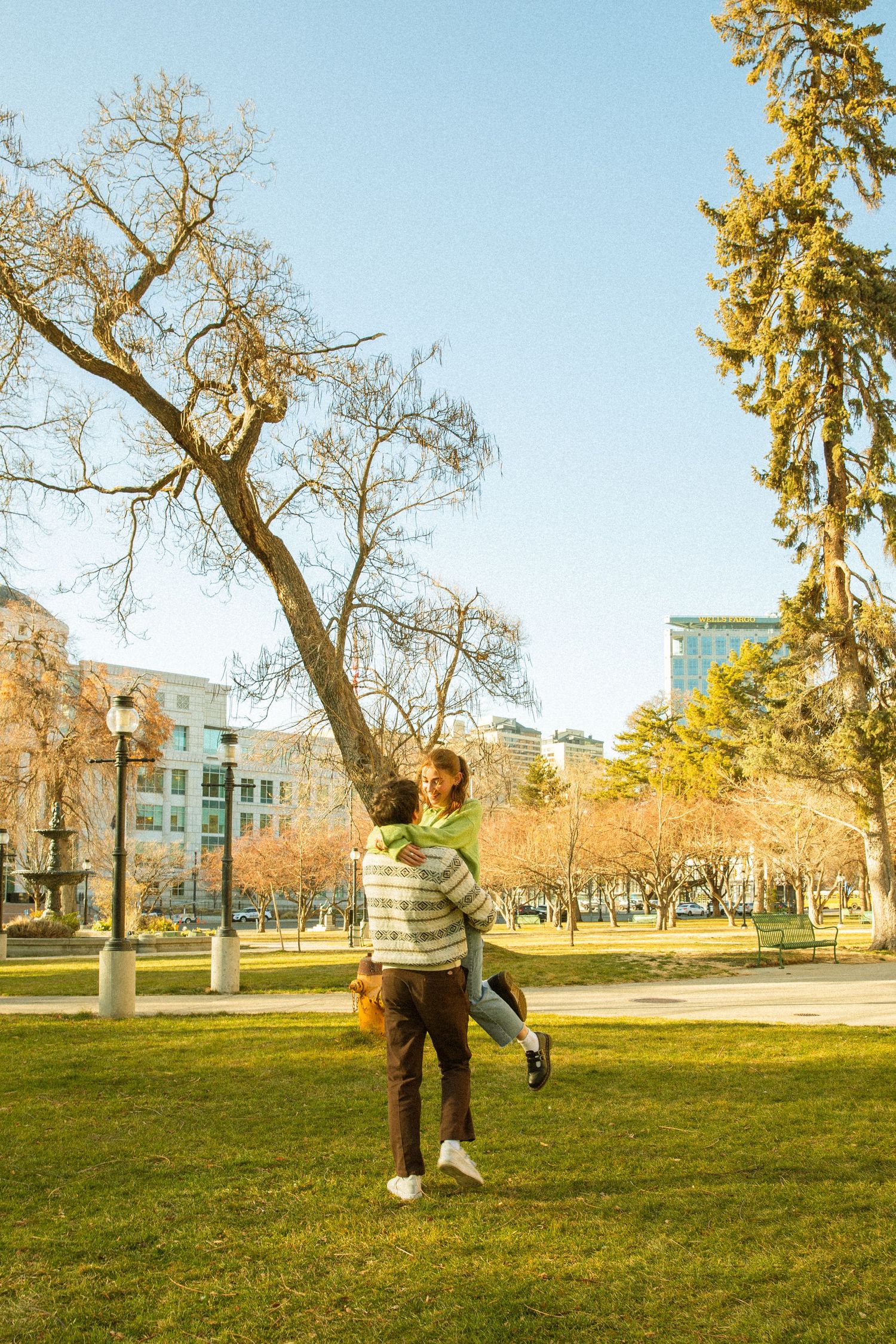 A person plays soccer on a grassy lawn surrounded by autumn trees and buildings on a sunny day.