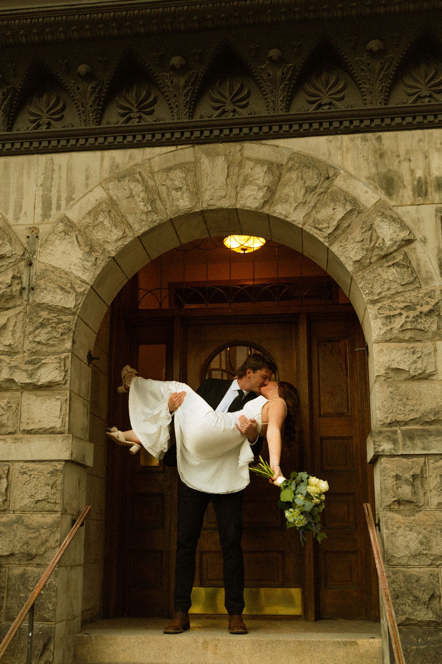 A romantic couple stands in an ornate stone archway entrance with wooden doors and decorative molding above.
