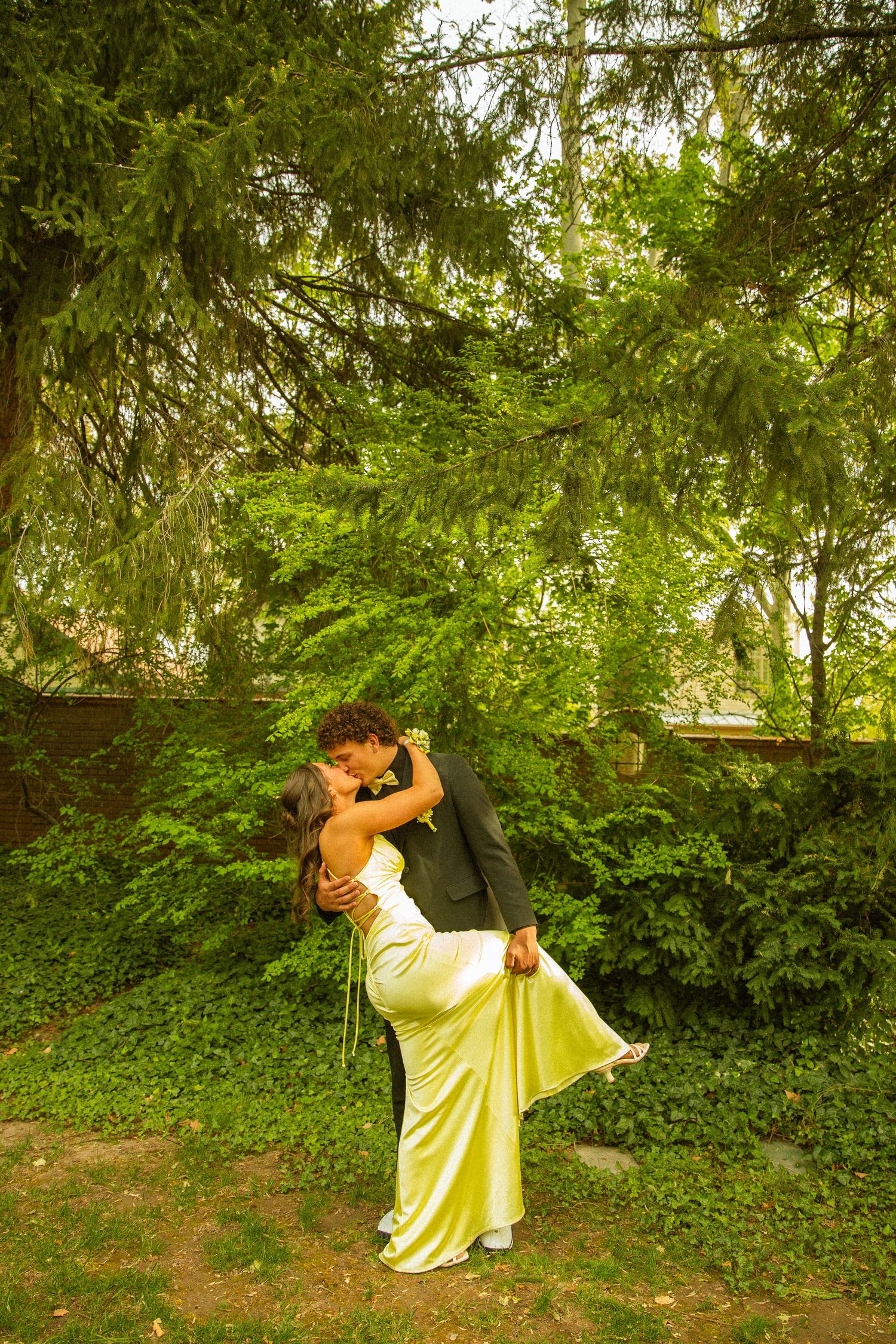 Couple sharing a romantic kiss in a lush green garden, woman in yellow satin gown, man in dark suit.