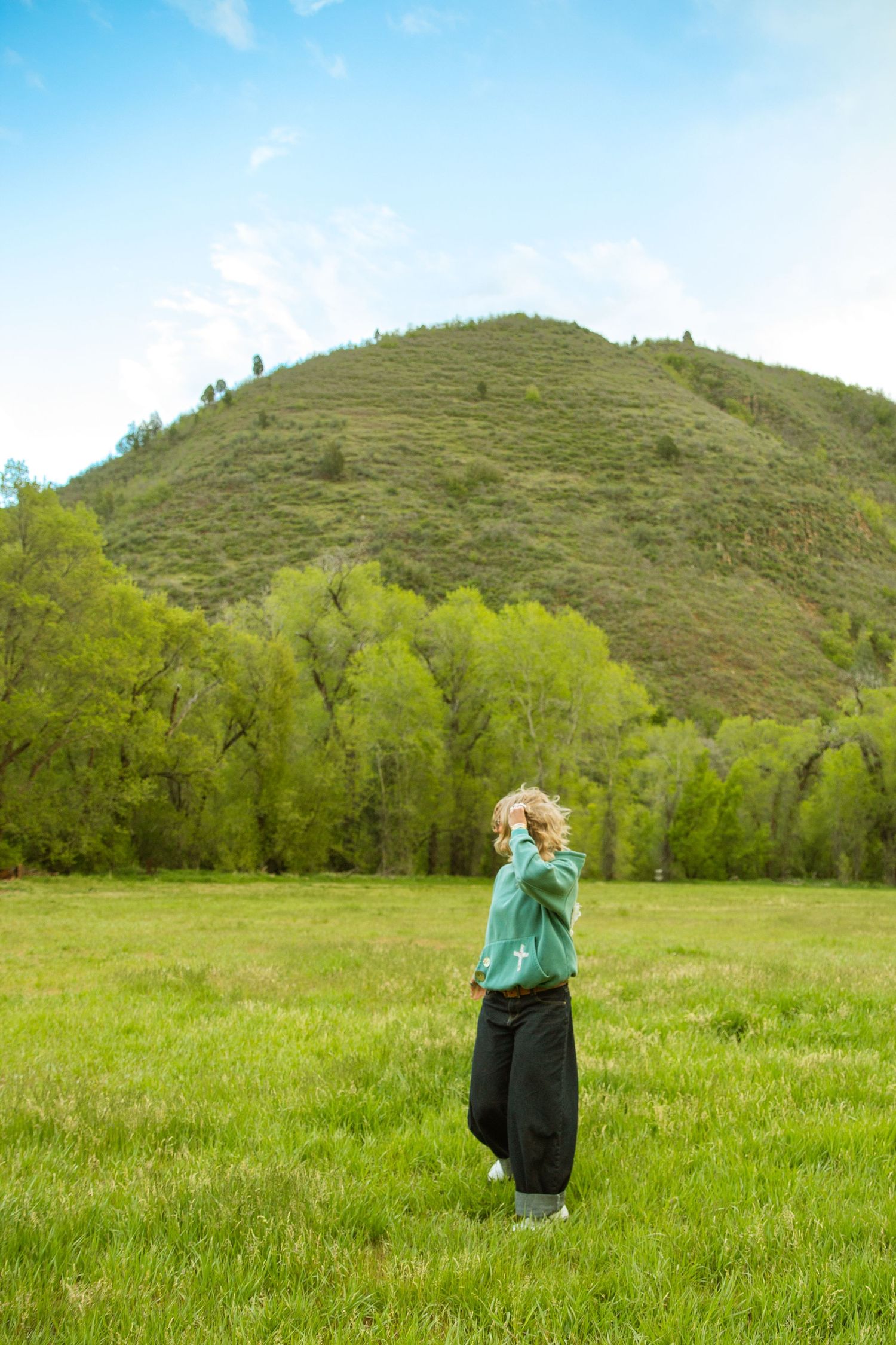 A young woman in a mint green hoodie stands in a lush green meadow with a tree-covered hill and blue sky behind her.