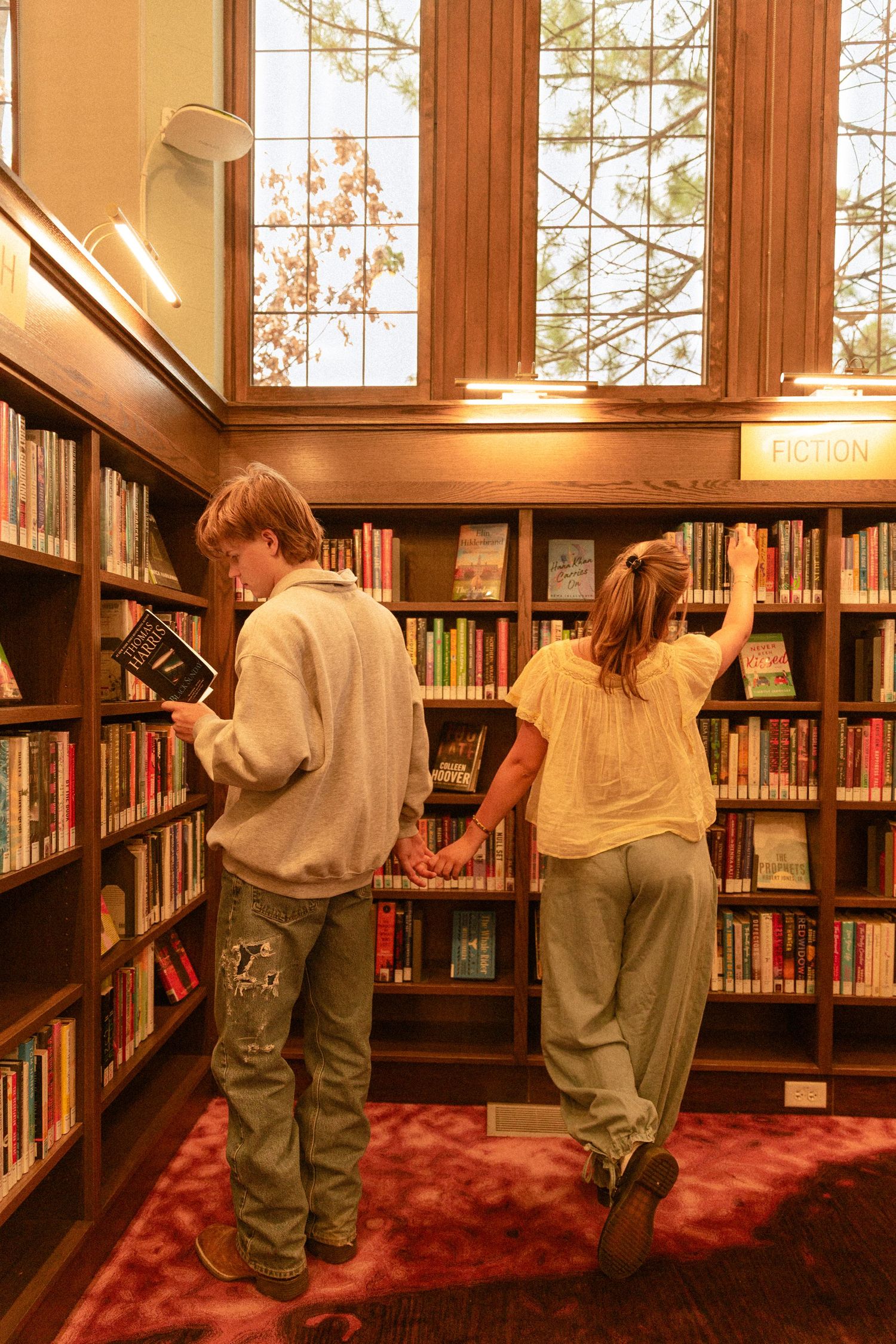 Multiple children browse books along wooden library shelves with sunlight streaming through windows above.