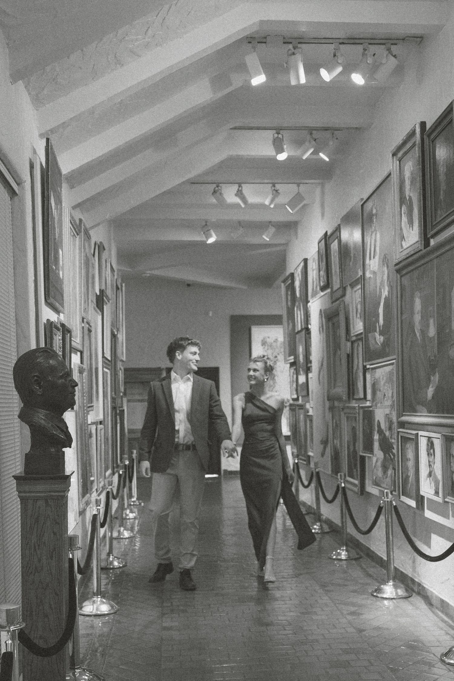 Vintage black and white photograph of a museum hallway with artwork displayed on walls and velvet ropes along the corridor.