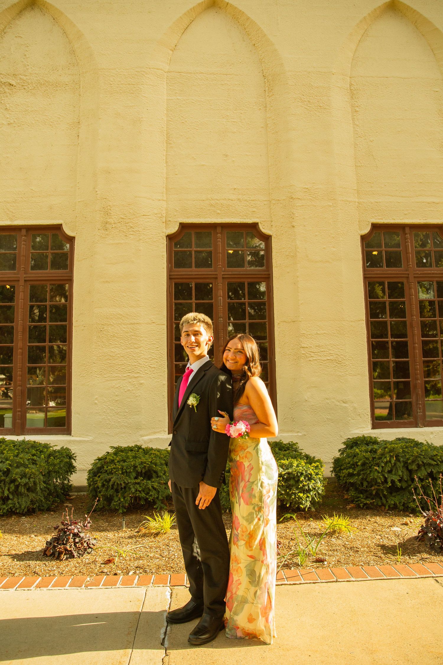 A young couple dressed in formal prom attire pose together outside an elegant building with arched windows.