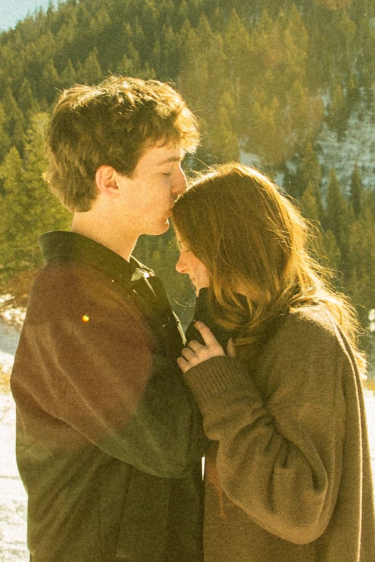 A couple shares an intimate moment in a snowy forest setting with golden sunlight filtering through the trees.