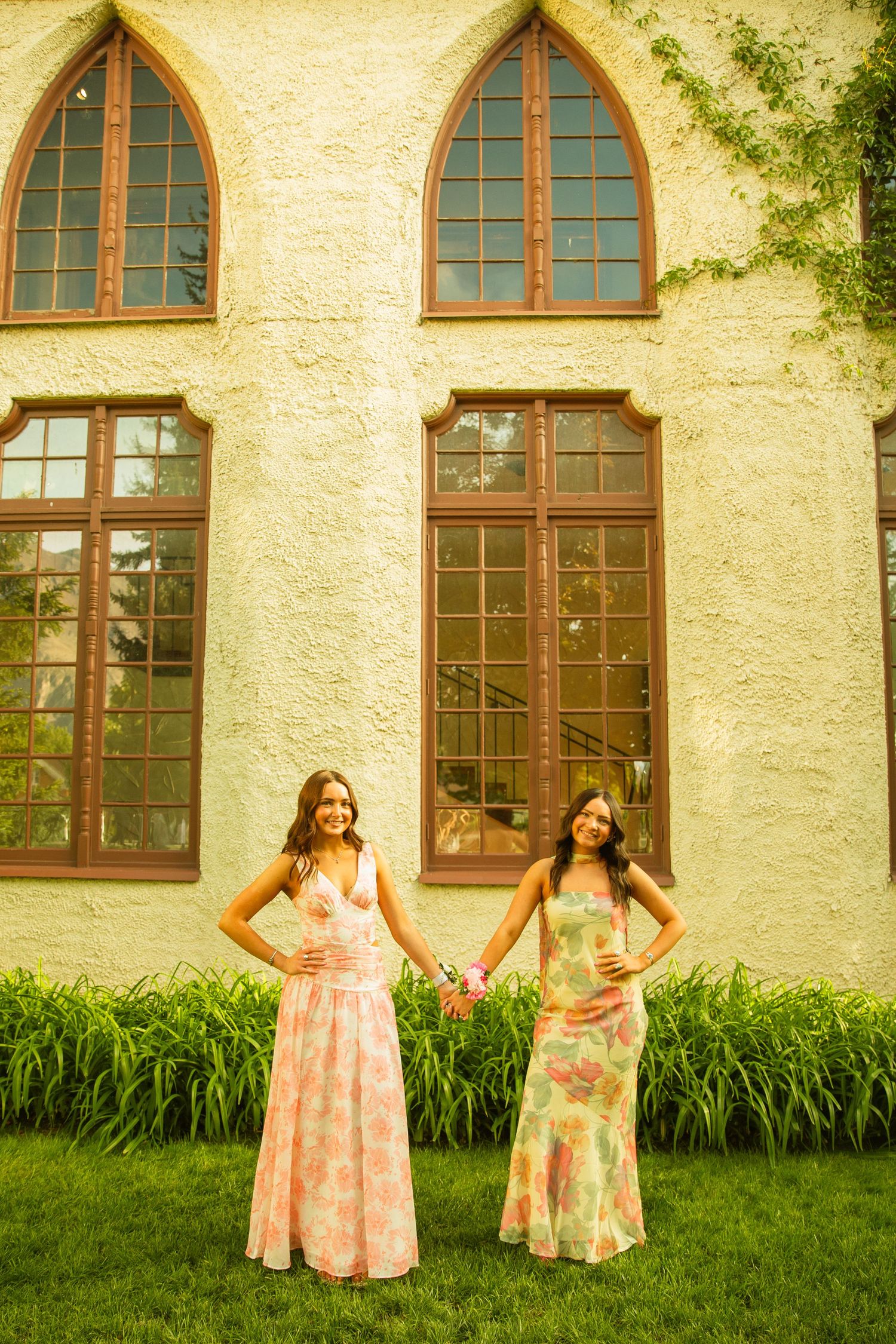 Two women in floral maxi dresses stand on green grass before a stone building with arched windows.