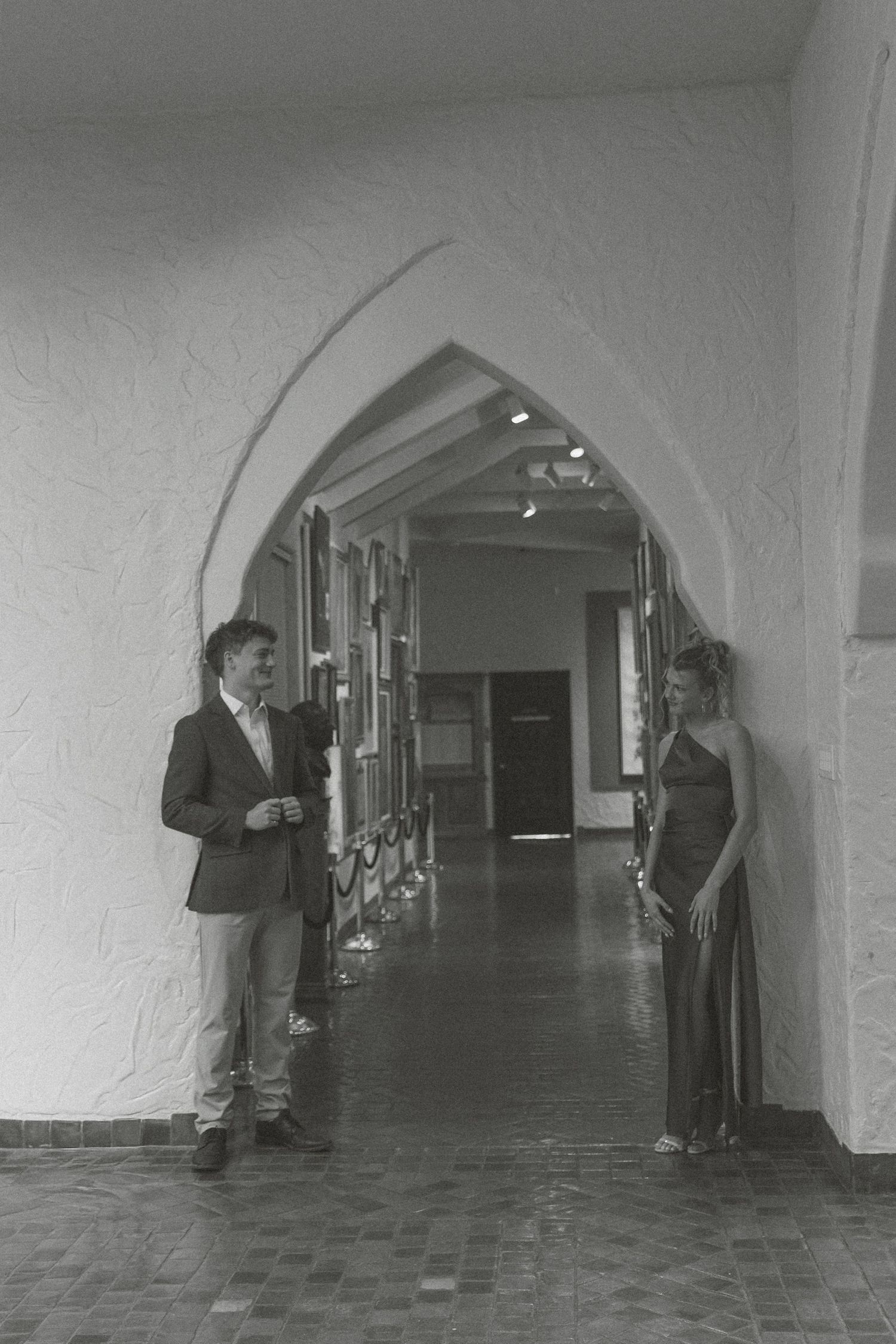 A black and white photo of two people standing in an arched hallway with textured walls and tiled floors.