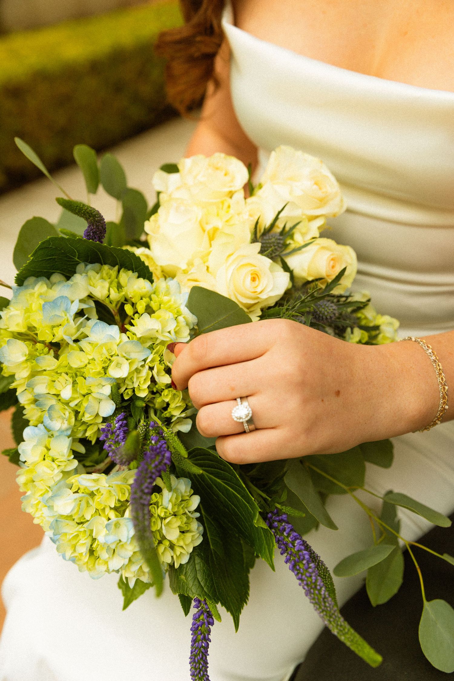 Close-up of a wedding bouquet featuring yellow roses, green hydrangeas, and purple flowers with a diamond ring.