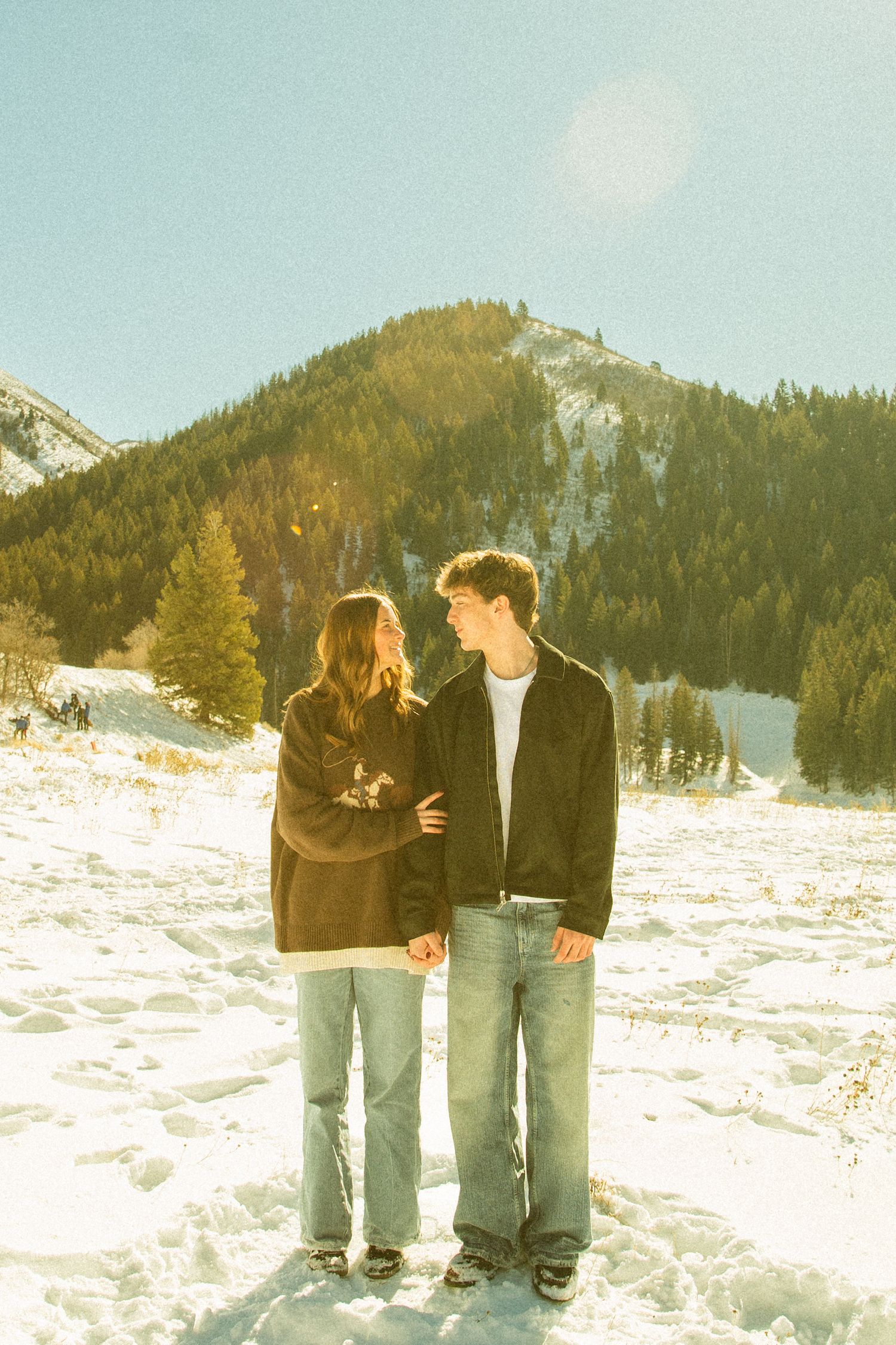 A couple stands together on snowy mountain terrain with evergreen forest and peaks in the background on a sunny day.