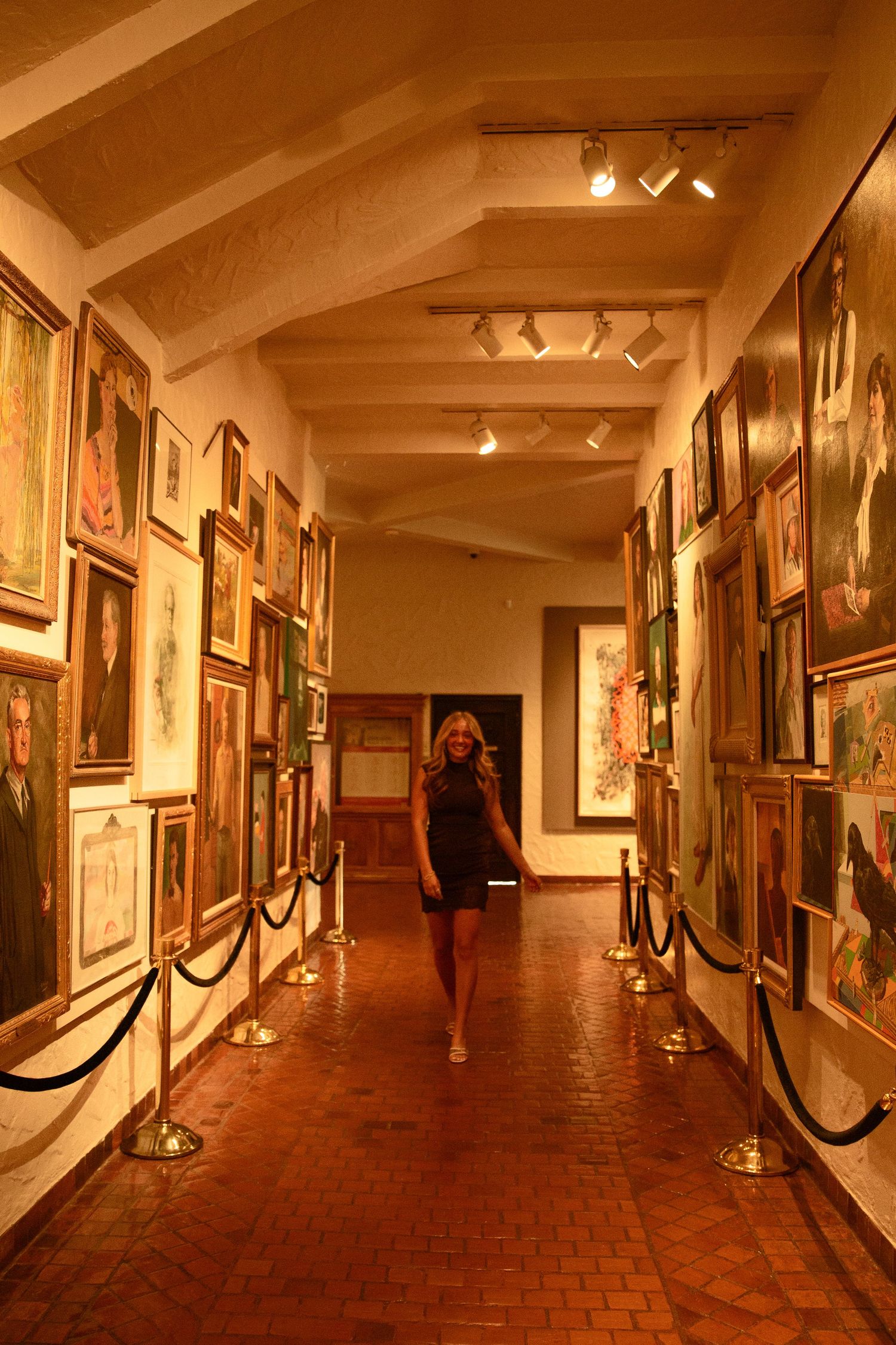 Woman walking through a narrow art gallery hallway lined with framed paintings and velvet rope barriers under warm lighting.
