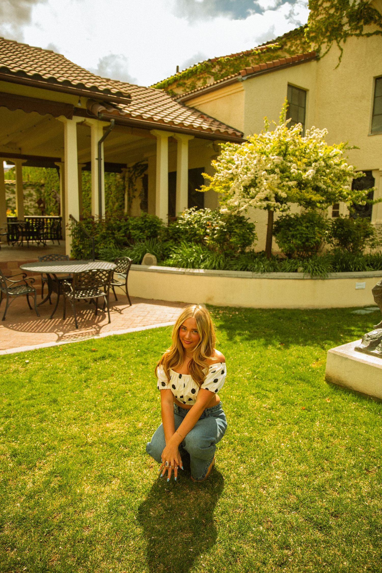 Young woman in polka dot top and jeans crouching on green lawn outside elegant Spanish-style home.