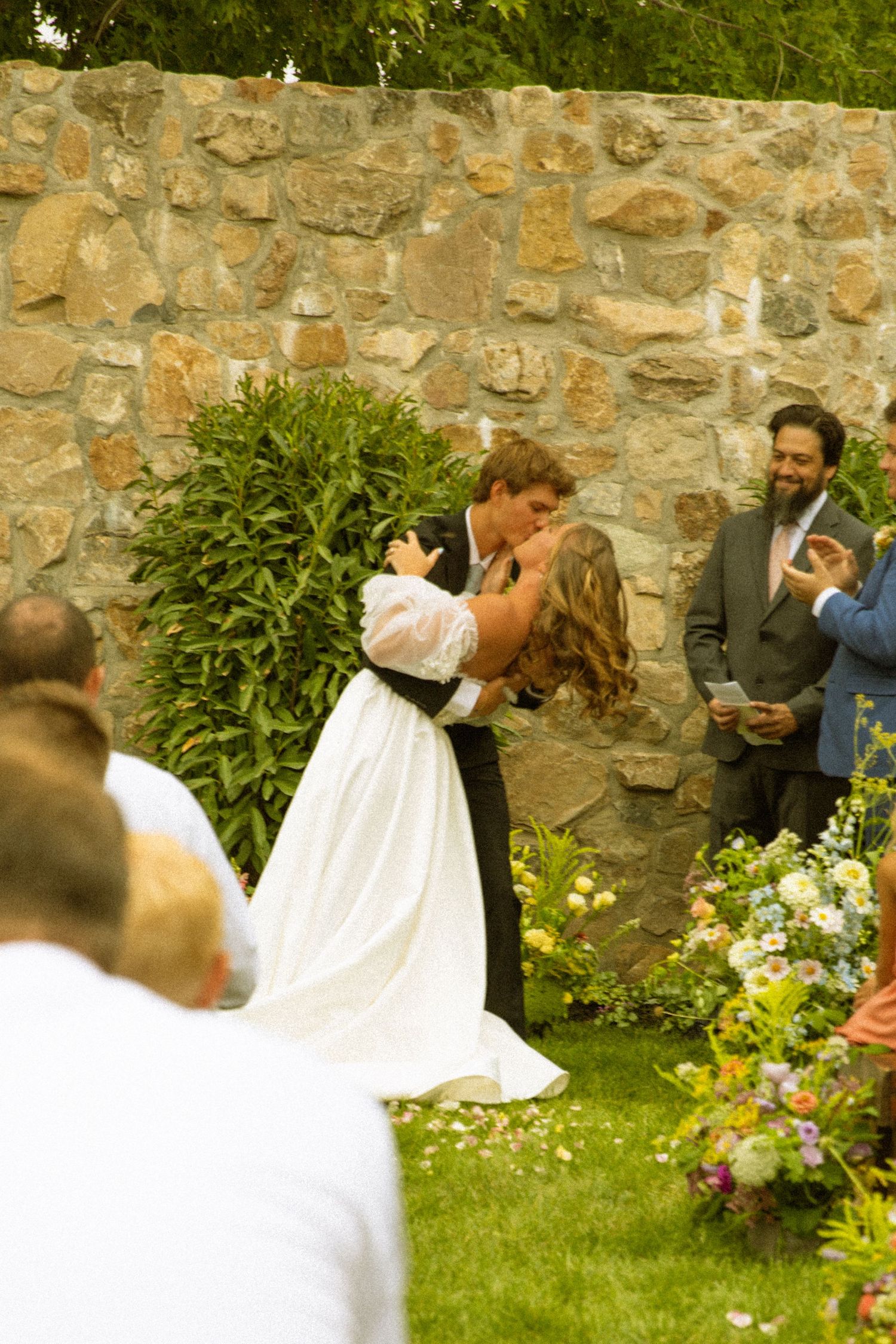 Wedding couple sharing a playful moment in a garden setting with stone wall backdrop.