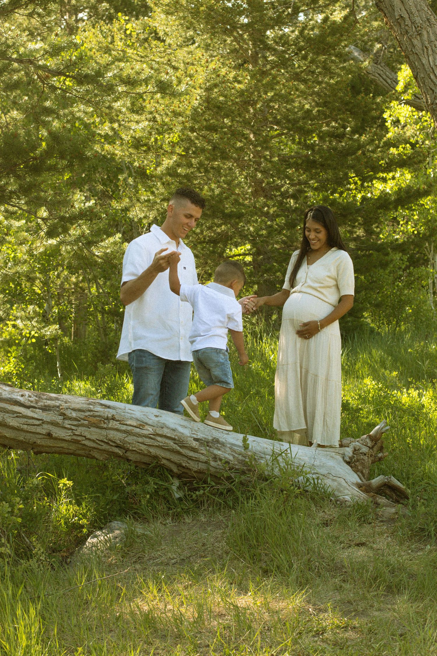 A couple in white clothing sits together on a fallen tree trunk in a sunlit wooded area during a photo session.