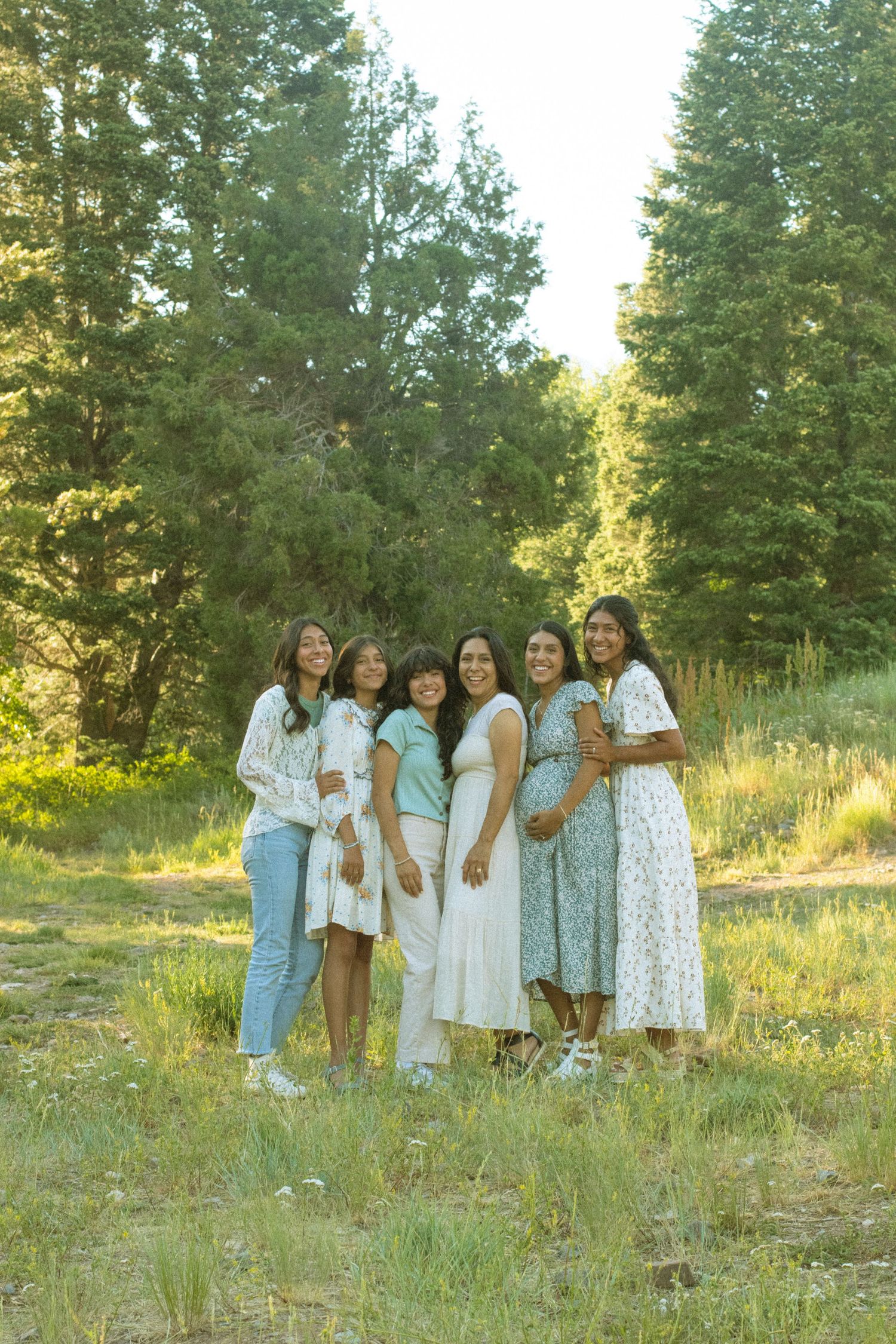 A group in light-colored dresses and denim stands together on a grassy hillside with evergreen trees in the background.