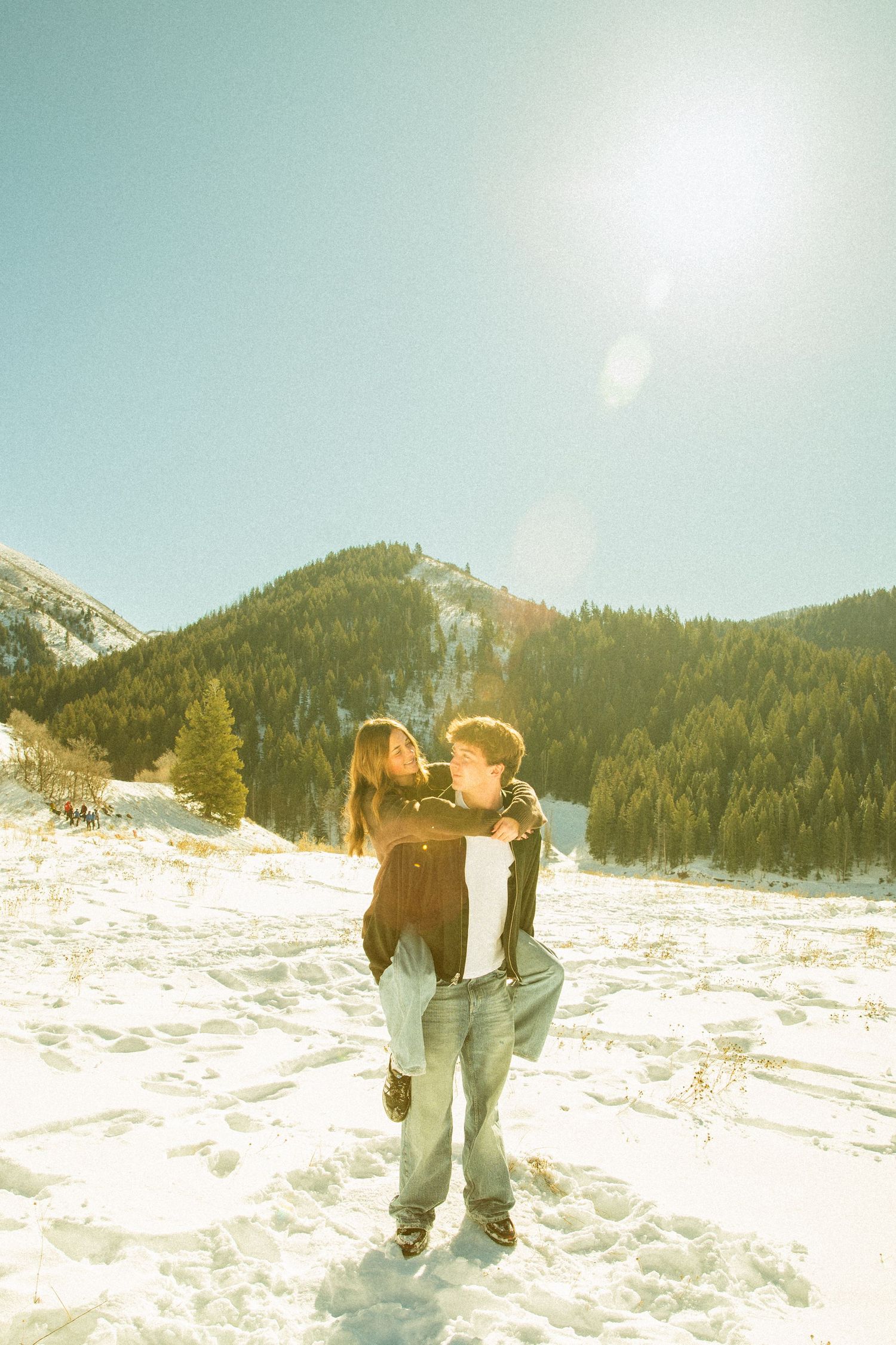 A couple shares a tender moment in the snowy mountains on a bright winter day with evergreen trees in the background.