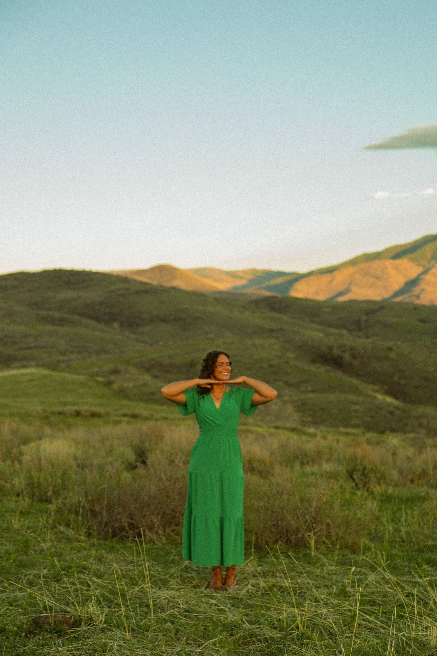 Woman in green midi dress standing in lush green mountain valley at golden hour sunset.